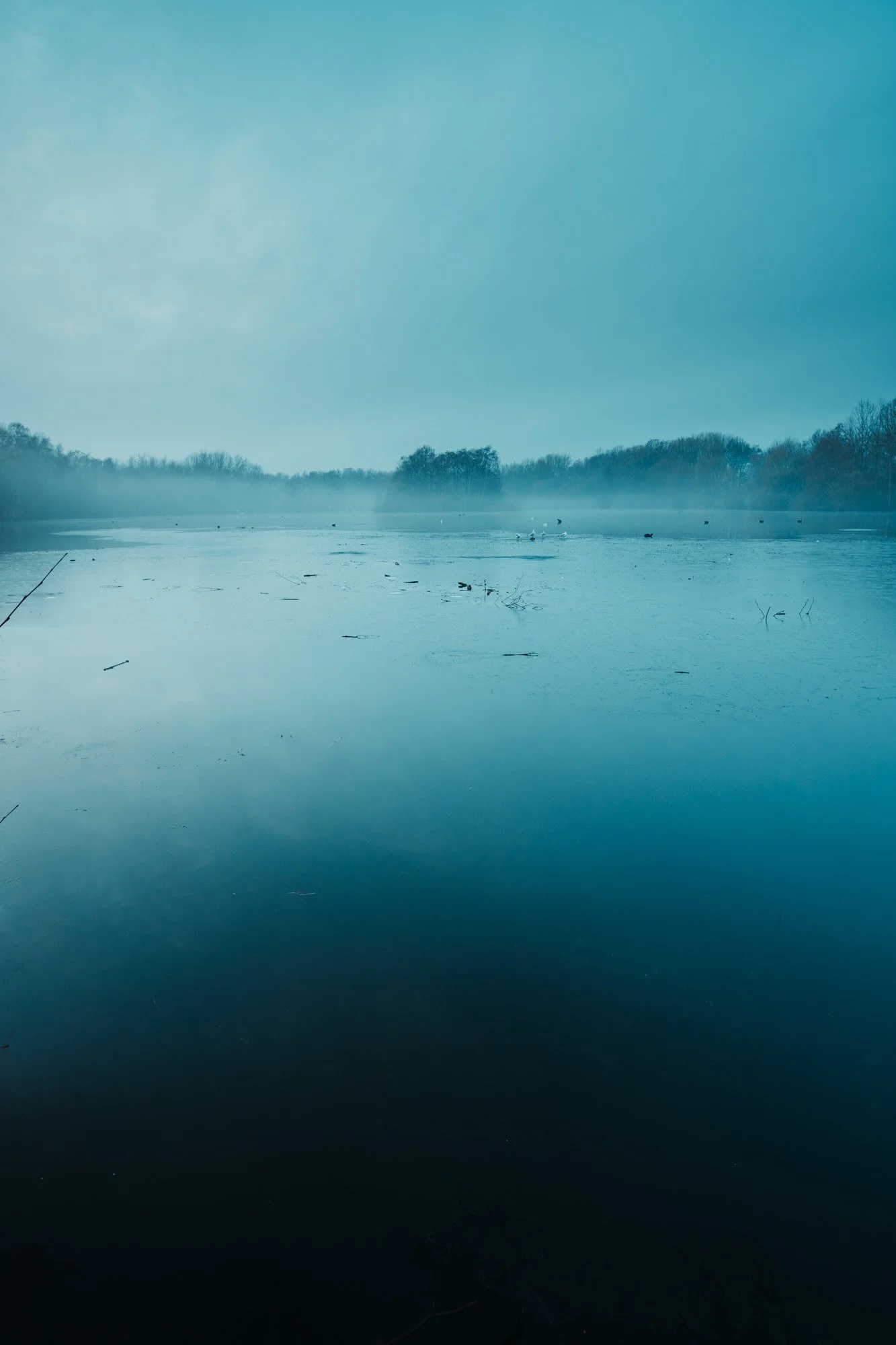 A misty lake with a calm, reflective surface, surrounded by trees in the distance the sky is mostly cloudy.