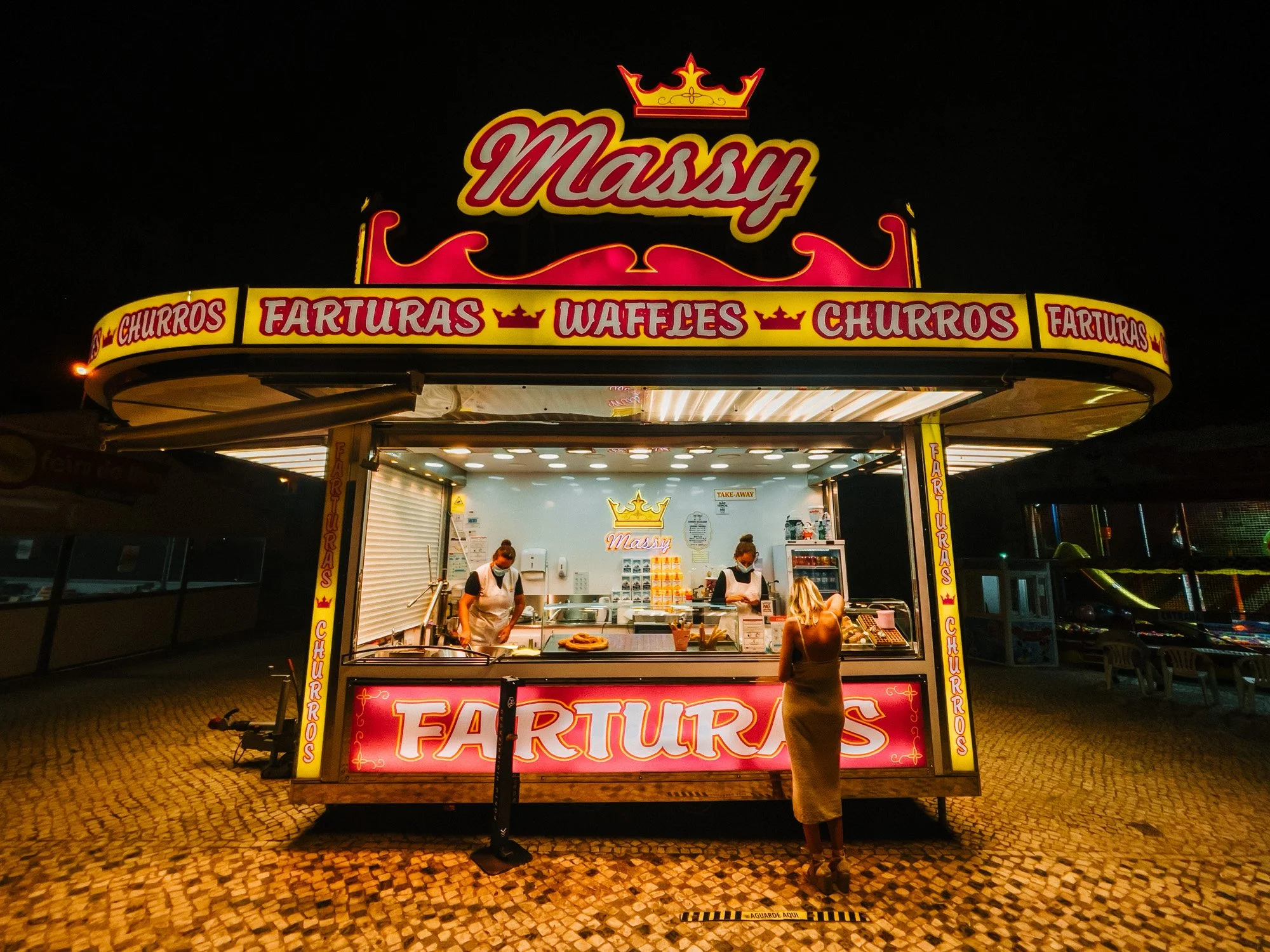 Night view of a brightly lit food stand named "Massy" selling churros, waffles, and fried treats, with a woman ordering from the stall.