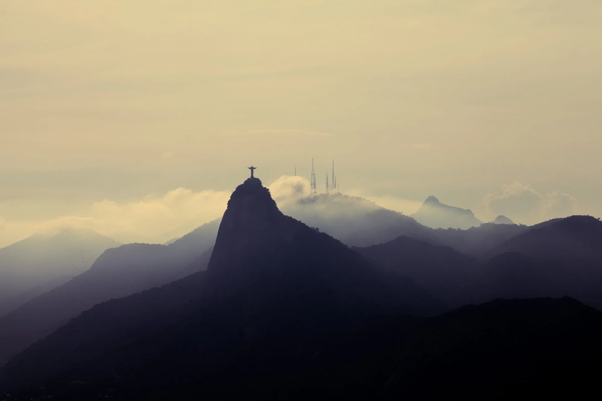 Silhouette of the Christ the Redeemer statue on Corcovado Mountain in Rio de Janeiro, with a foggy sky and distant mountain peaks.