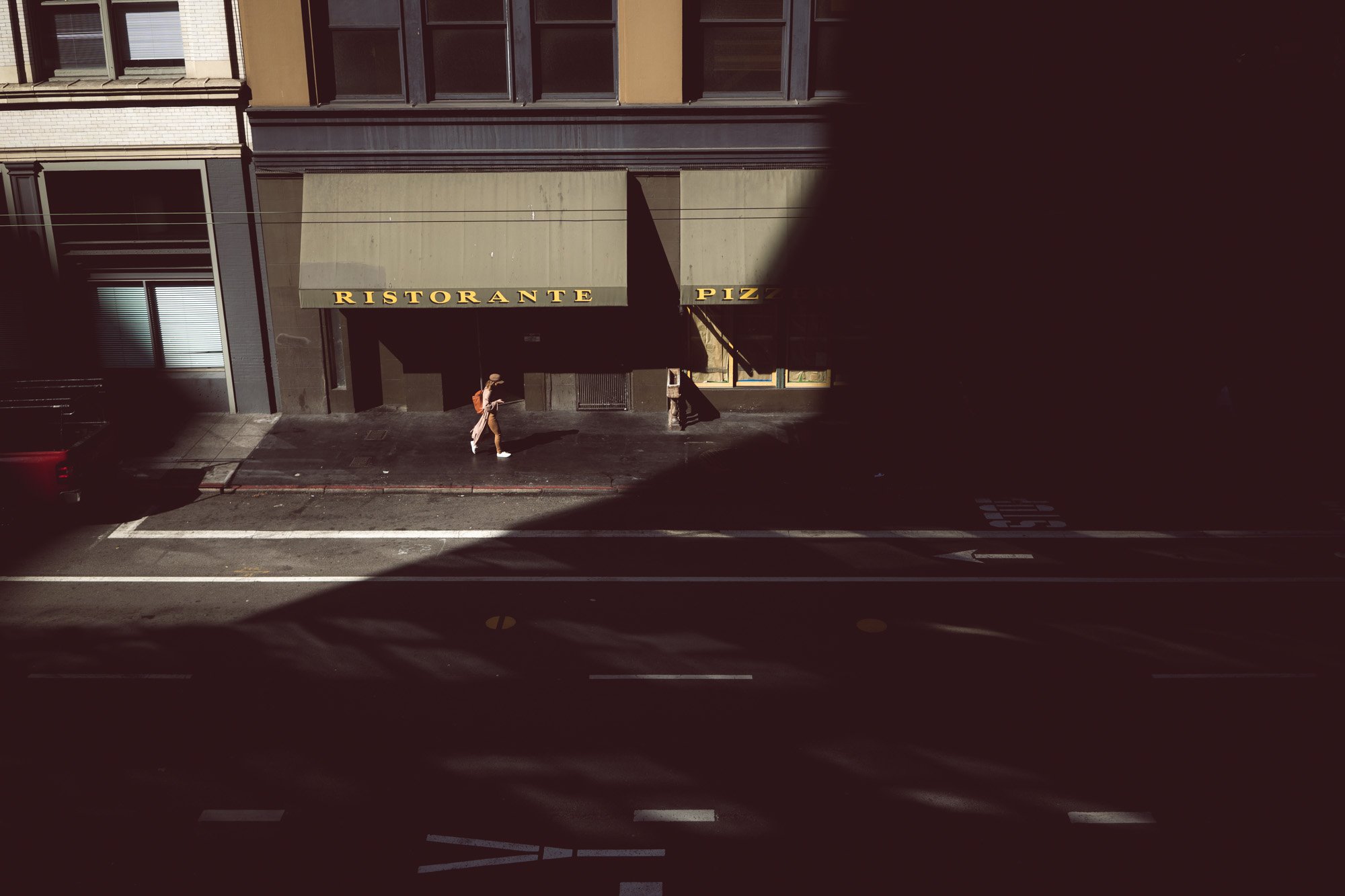 A street view with a building that has a sign reading 'Ristorante Pizzeria' above the entrance. Pedestrians walk on the sidewalk, and shadows cast by nearby structures are visible on the ground.