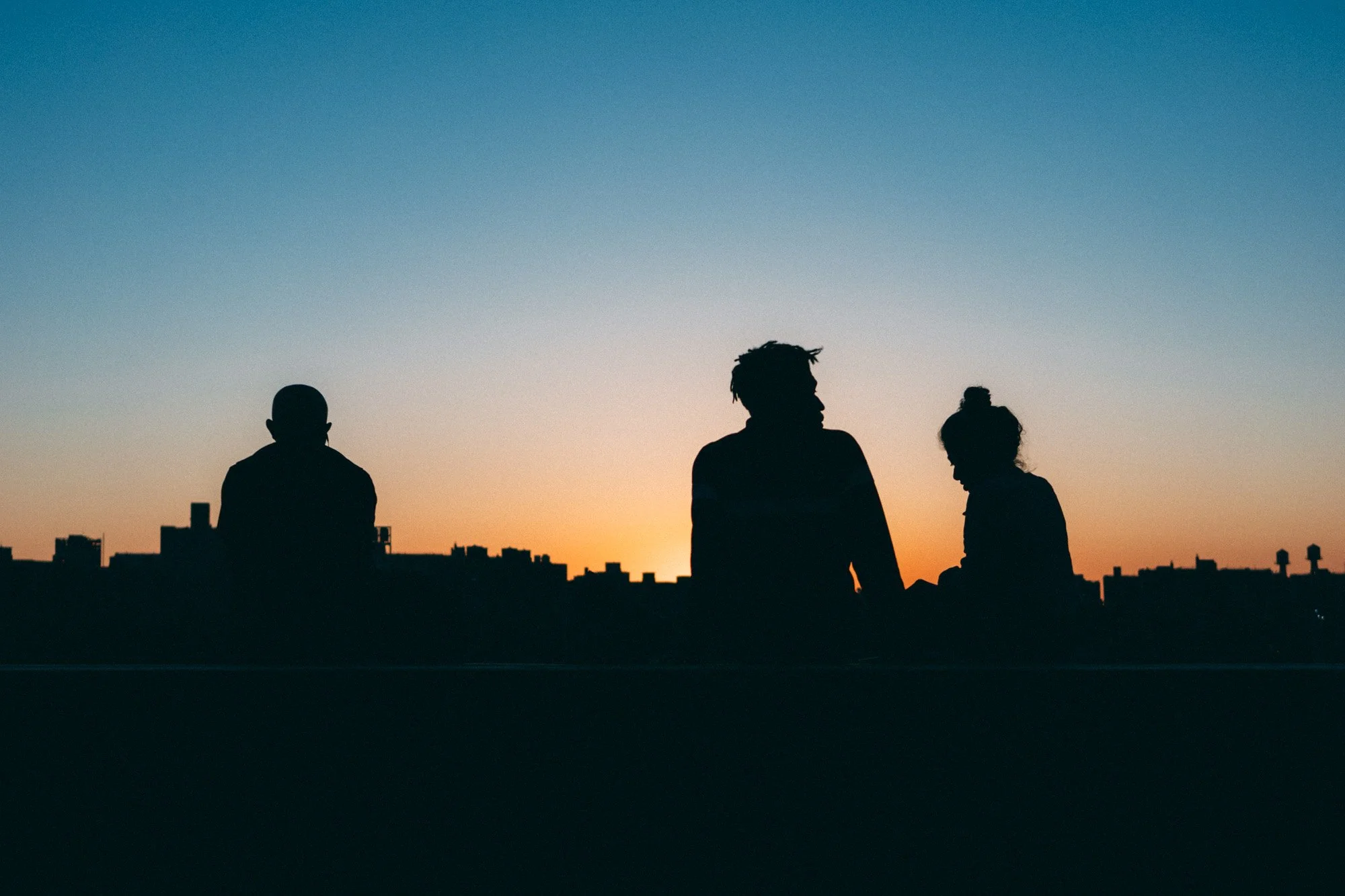 Silhouettes of three people sitting on a rooftop during sunset with city buildings in the background.