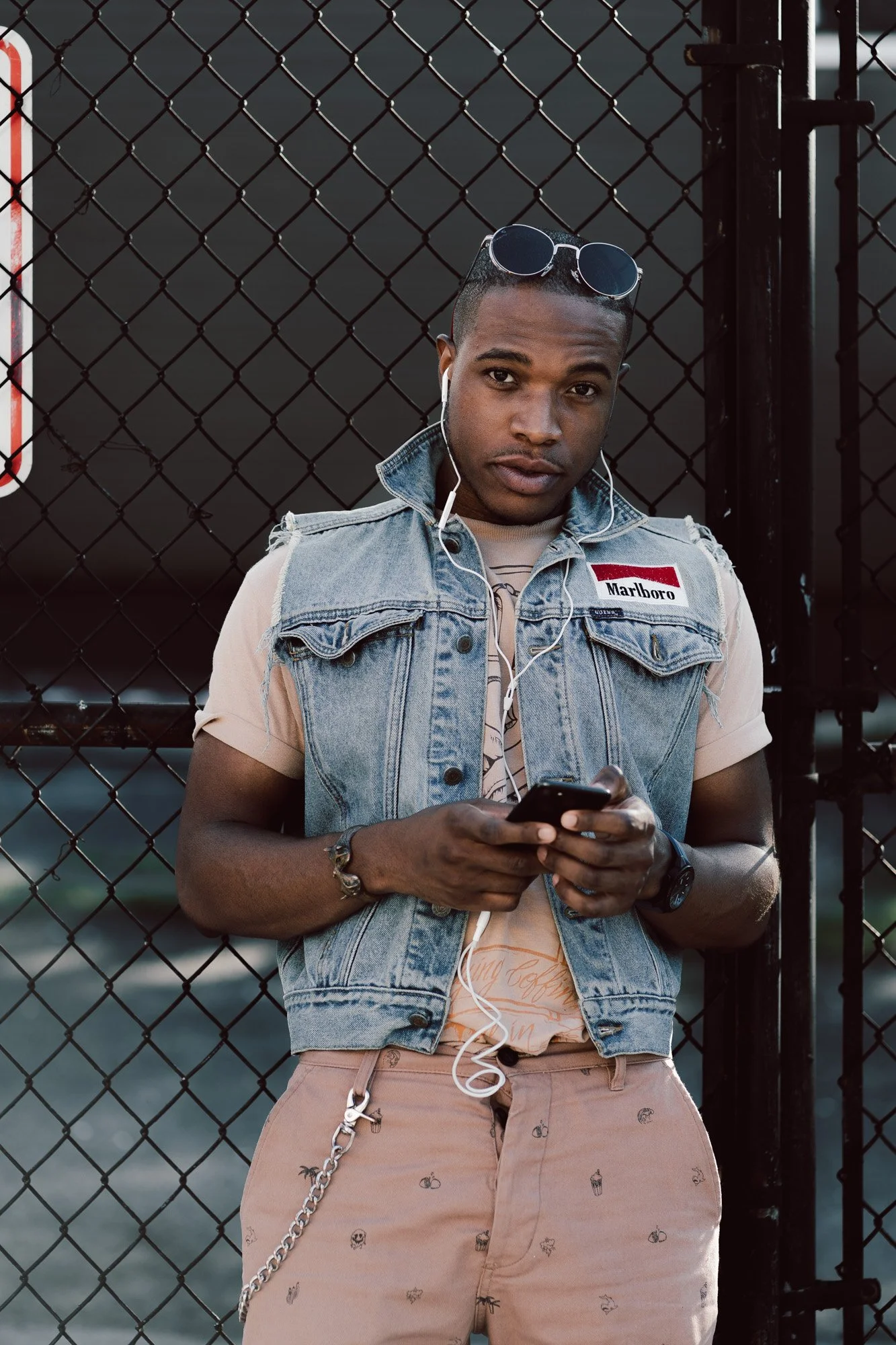 A young man with dark skin wearing a sleeveless denim vest with a Marlboro patch, peach-colored pants with small patterns, listening to music with white earphones, holding a smartphone, standing in front of a chain-link fence.