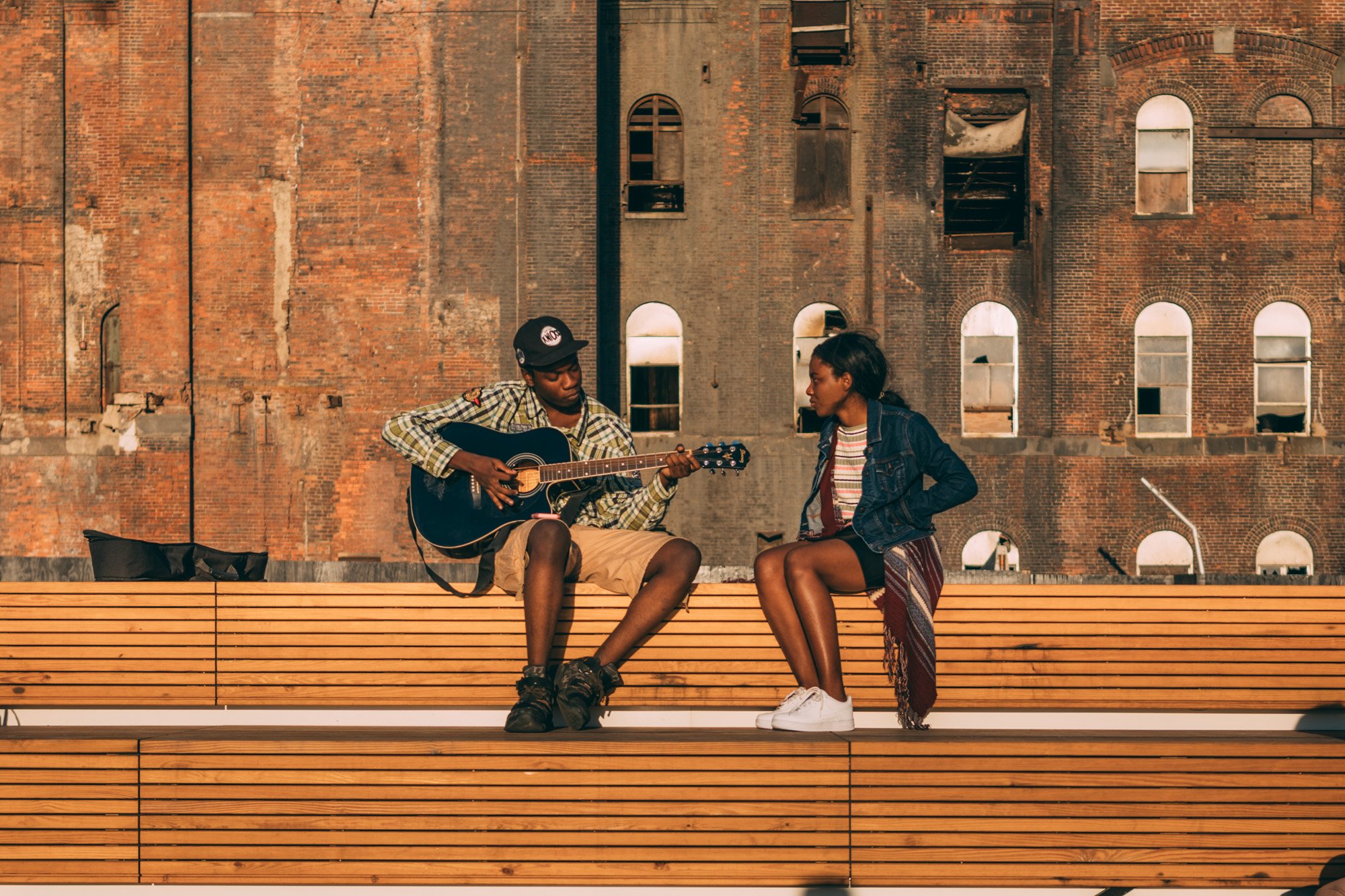 A young man playing guitar and a young woman sitting nearby on a wooden bench, engaging in conversation, with a brick building in the background.