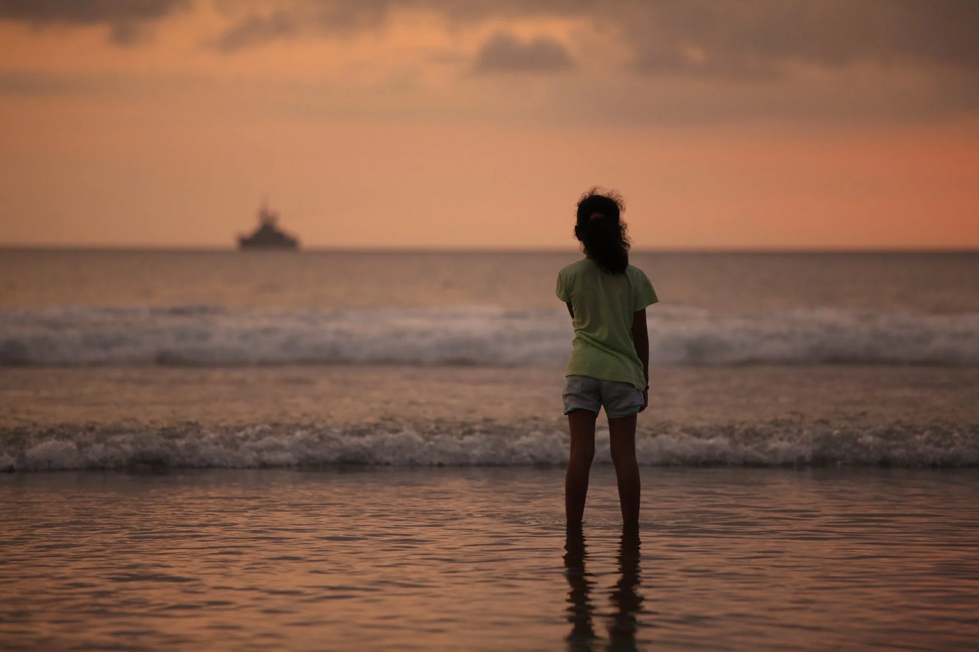 A girl stands in shallow water on the beach at sunset, with her back to the camera, looking out at the sea and a distant ship on the horizon.