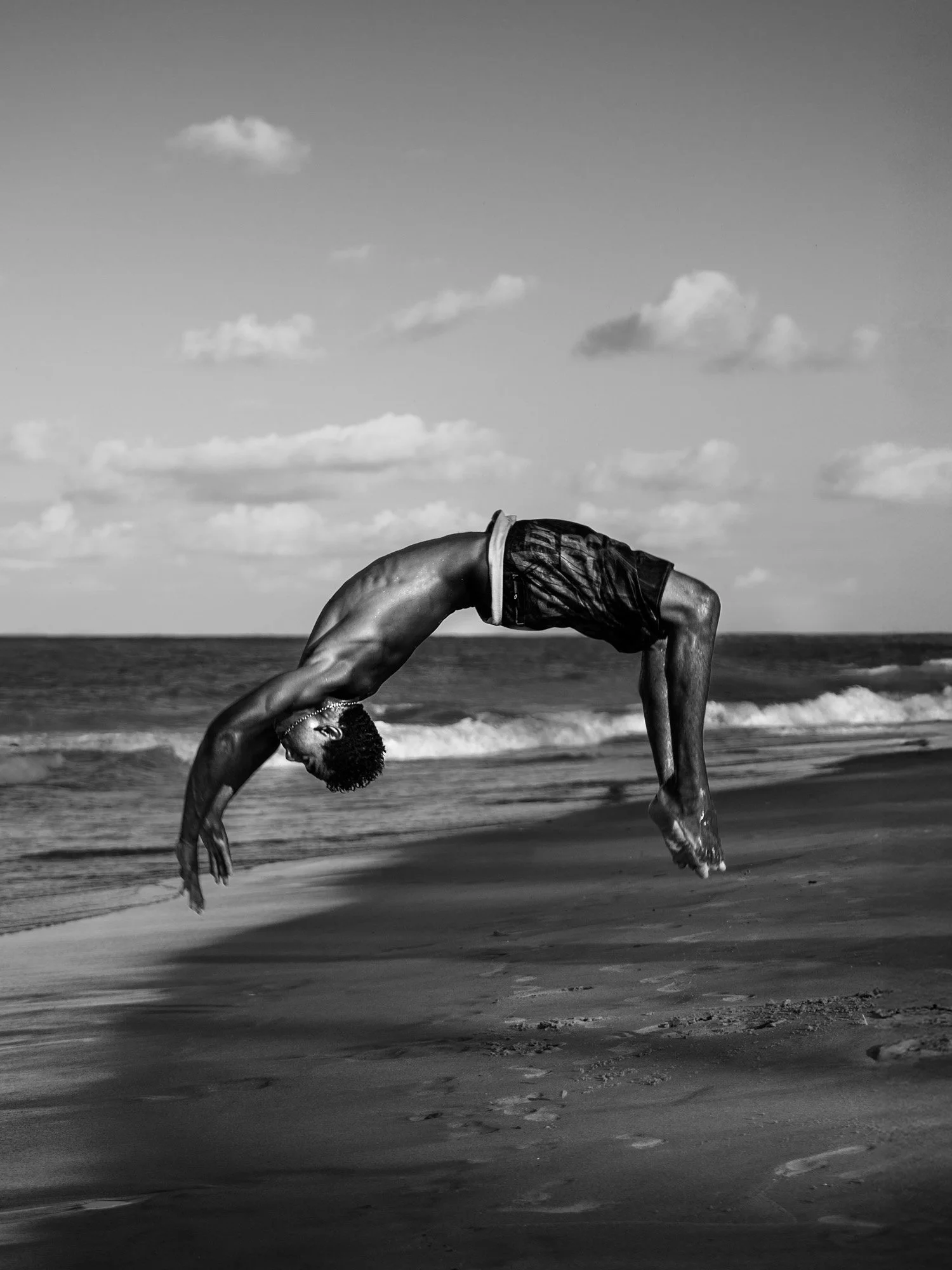 A person performing a backflip on the beach during the daytime, with ocean waves and partly cloudy sky in the background.