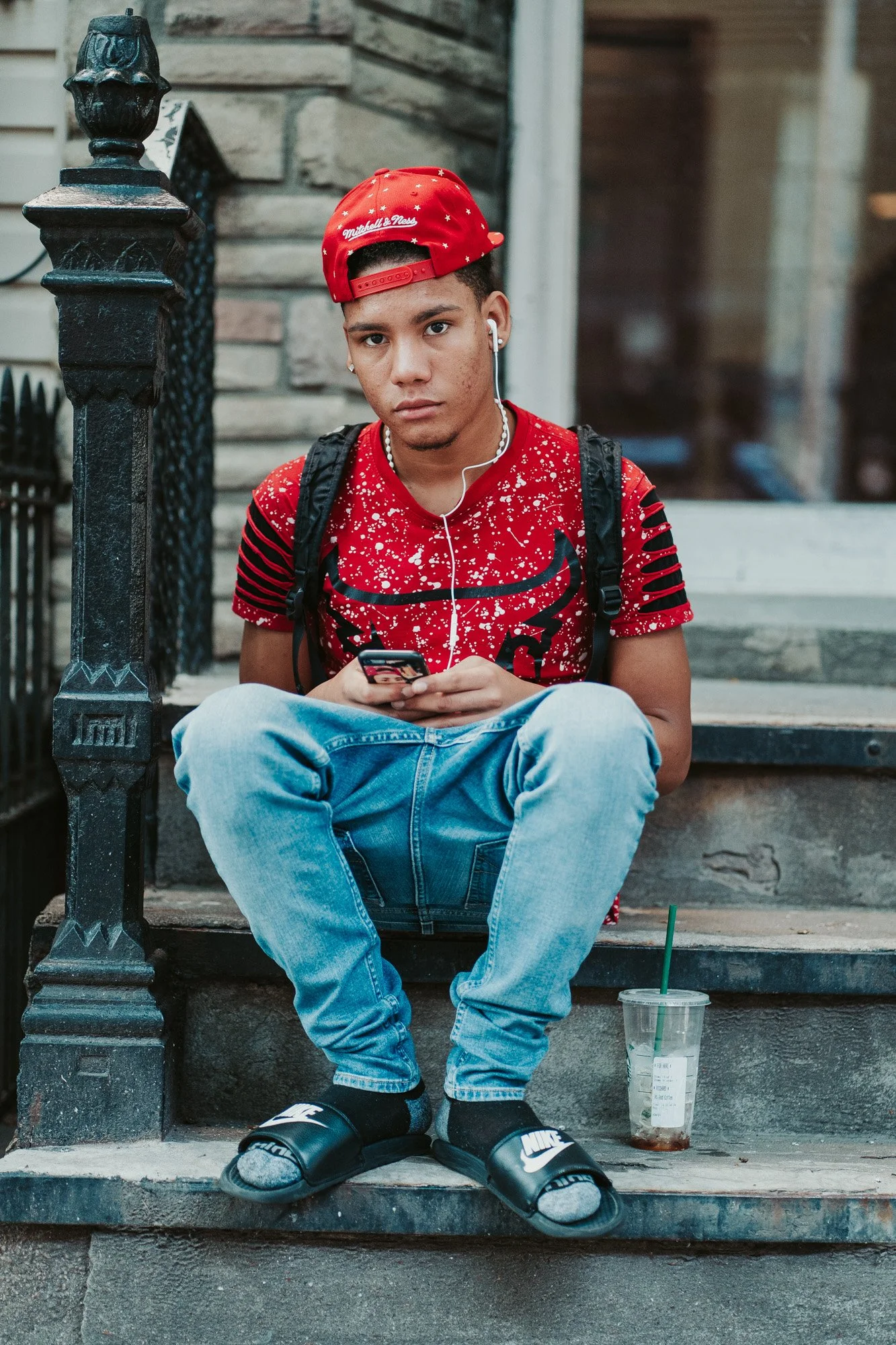 A young man sitting on the steps outside a building, wearing a red cap, a red graffiti shirt, light blue jeans, and black Nike slides with socks. He is holding a smartphone and has earphones in. There is a partially empty iced drink cup on the step next to him.