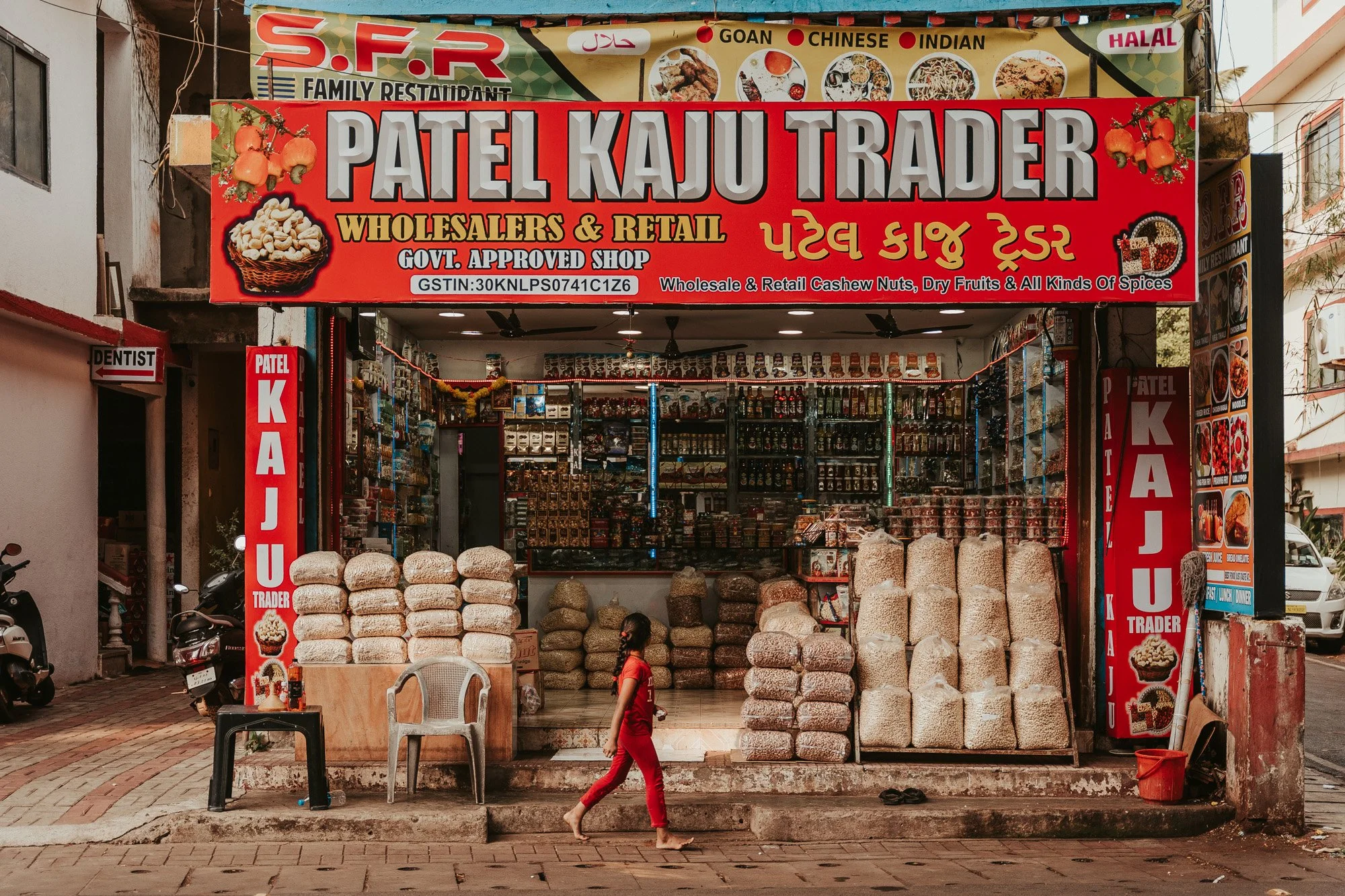 A street view of Patel Kaju Trader store selling cashew nuts, dry fruits, spices, and other products. The store has large red and yellow signs with shop name and offerings in English and Gujarati. There are bags of cashew nuts outside, and a girl walking by barefoot. The shop is decorated with garlic and chili strings, and has a colorful display of products inside.