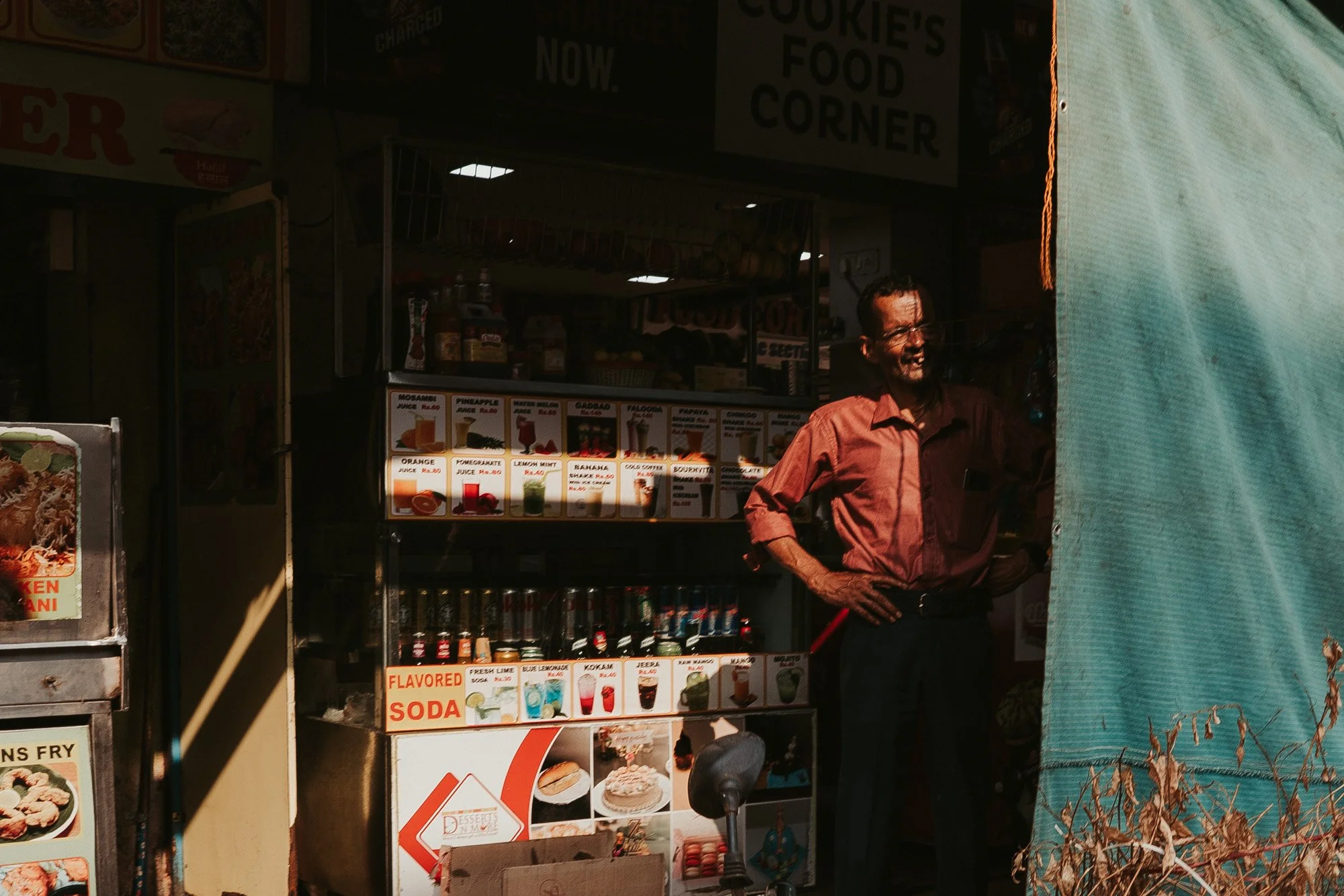 A man standing inside a food stall, smiling, with various drink and food menus displayed behind him, partially obscured by a blue fabric curtain on the right side.