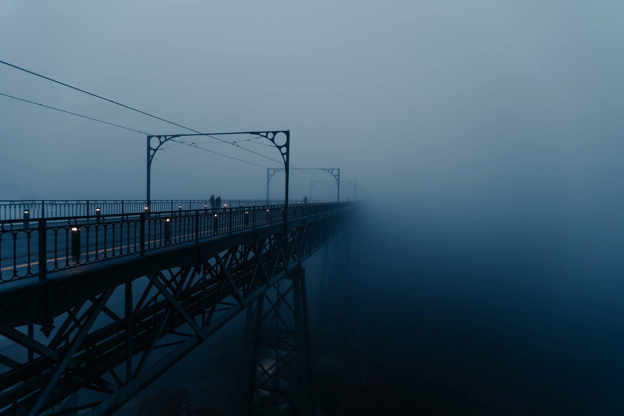 A foggy view of a steel bridge with a few people walking on it, faintly visible in dense fog with a dark, mysterious atmosphere.
