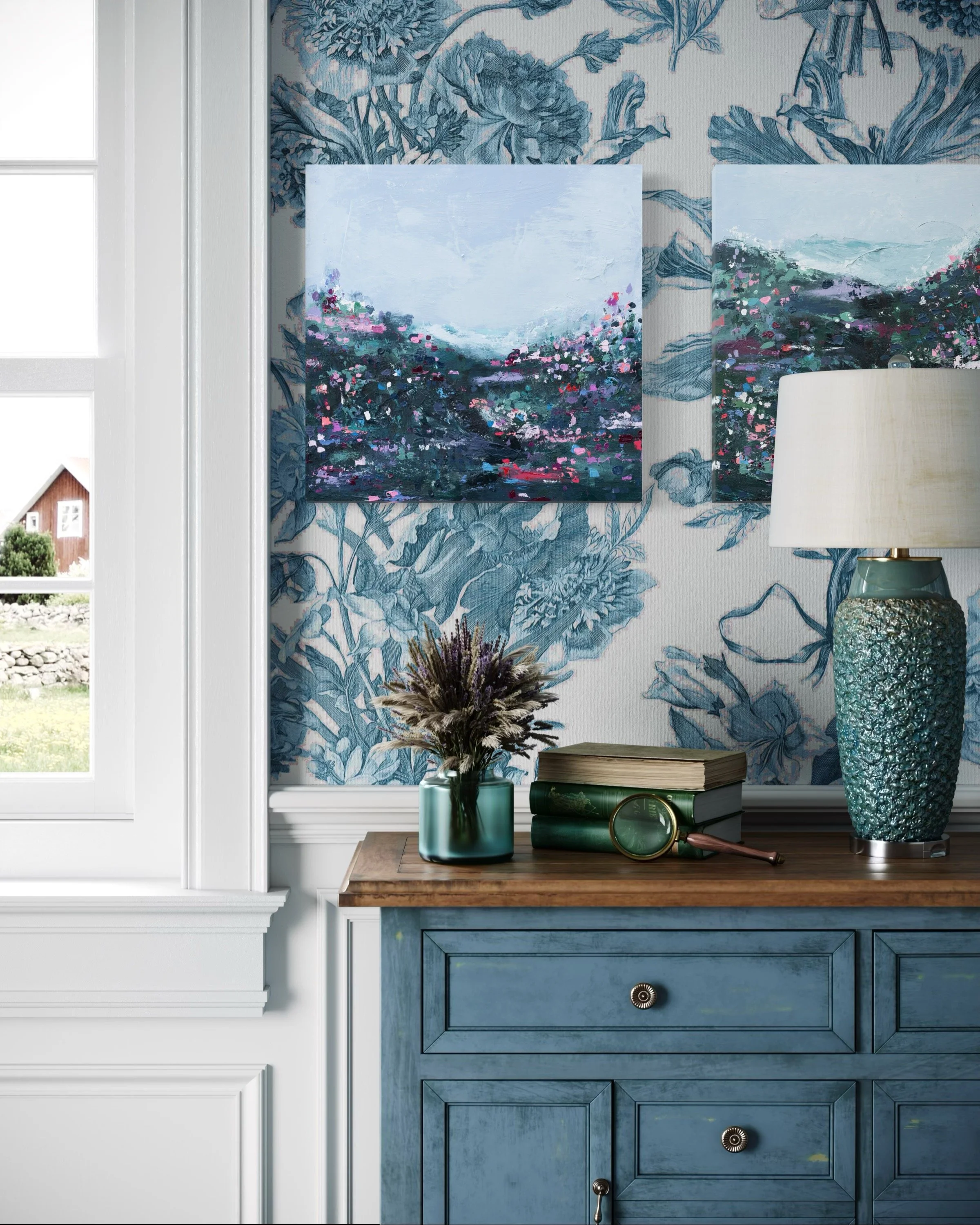 Interior shot of a blue dresser with decorative items including a book, a magnifying glass, a vase with dried flowers, and a lamp. The wall behind has floral wallpaper and two landscape paintings by Jocelyn Elizabeth.