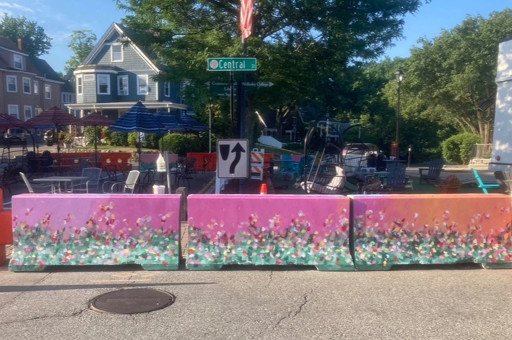 Colorful flower-patterned barriers blocking part of the street, with outdoor chairs and umbrellas behind them, and a street sign for Central Street visible in a residential neighborhood.