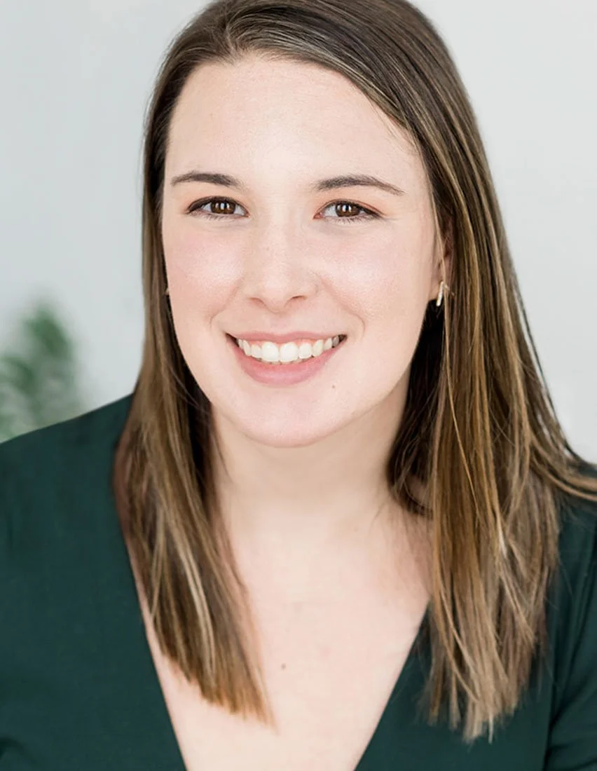 A smiling woman with light skin, brown hair, and brown eyes, wearing a dark green top and small hoop earrings, posed against a plain light background.