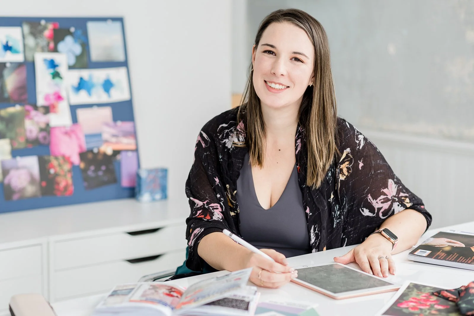 A woman sitting at a desk with magazines, a tablet, and a magazine in front of her, smiling at the camera. There is a board with photos and illustrations on the wall behind her.