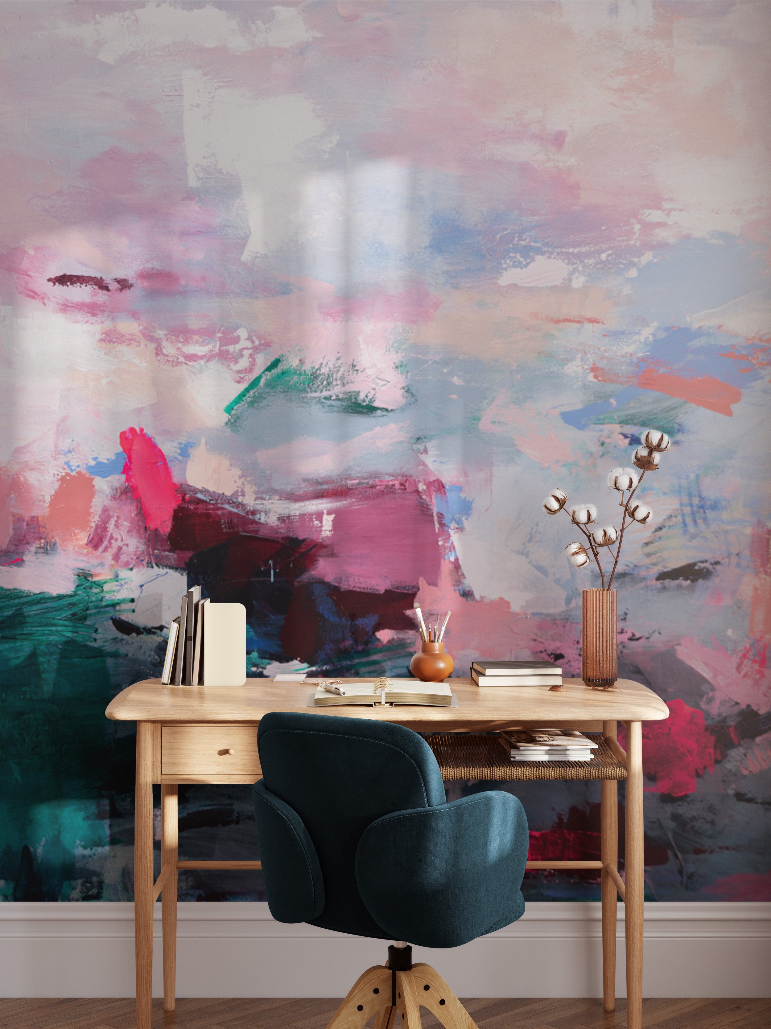 Interior of a study space with a wooden desk, a dark green desk chair, and an abstract colorful mural wall with pink, green, purple, and blue brushstrokes, decorated with books, a vase with cotton flowers, and stationery.