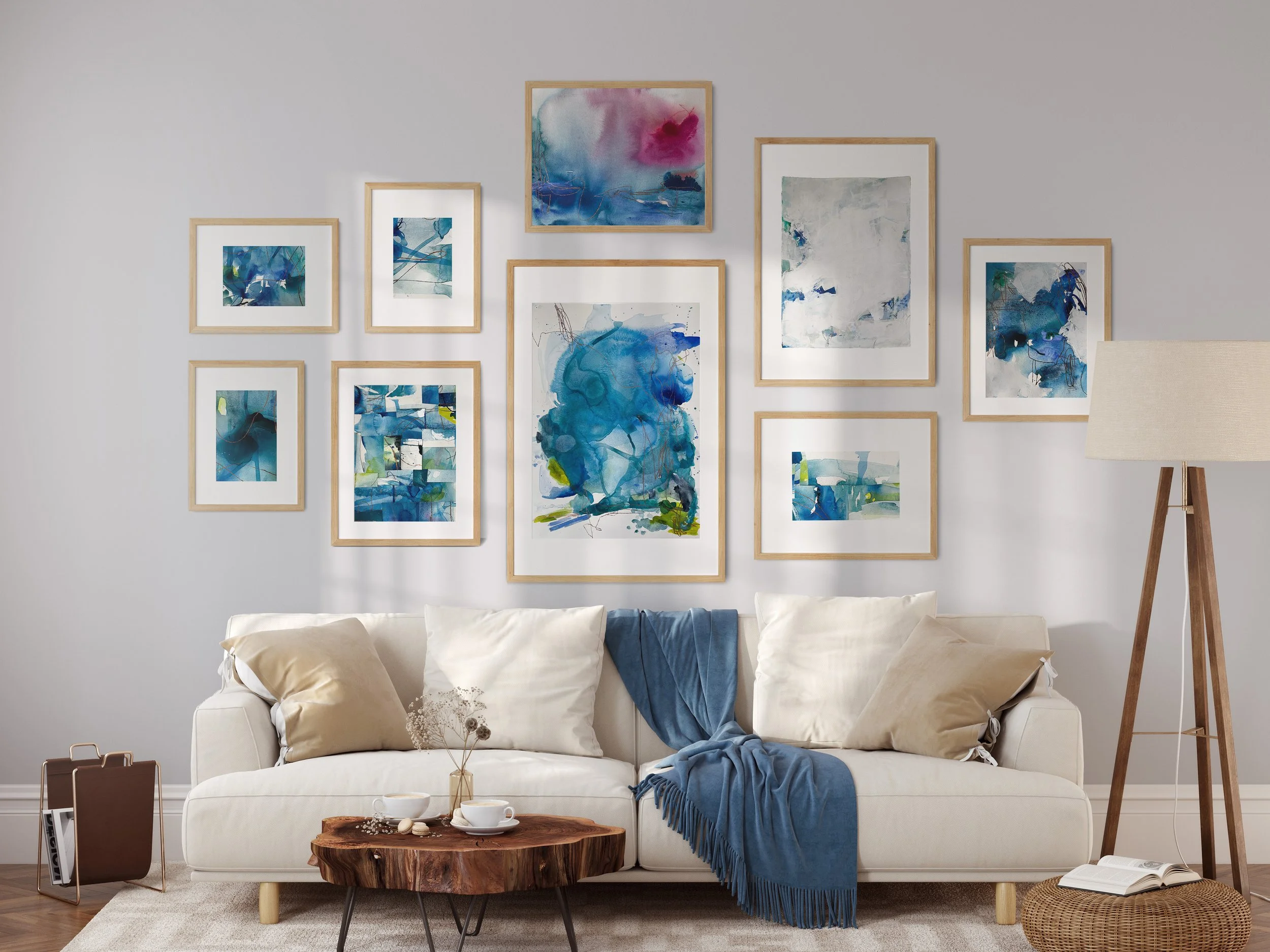 Living room with a white couch, beige and cream pillows, a blue throw blanket, a wooden coffee table with teacups, a small vase, and decorative items, framed abstract blue and white artwork on the wall, a floor lamp with beige shade, a small side tab