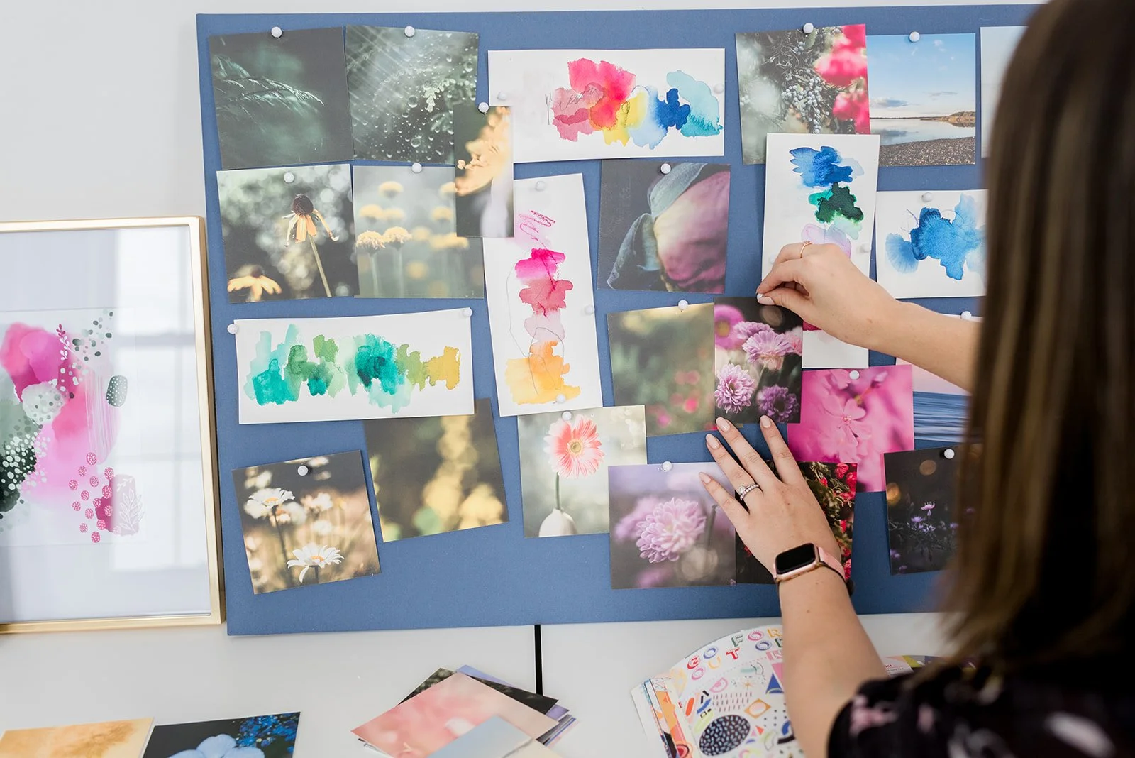 Person attaching pink flower photographs and watercolor artworks to a blue bulletin board, with print materials on the table below.