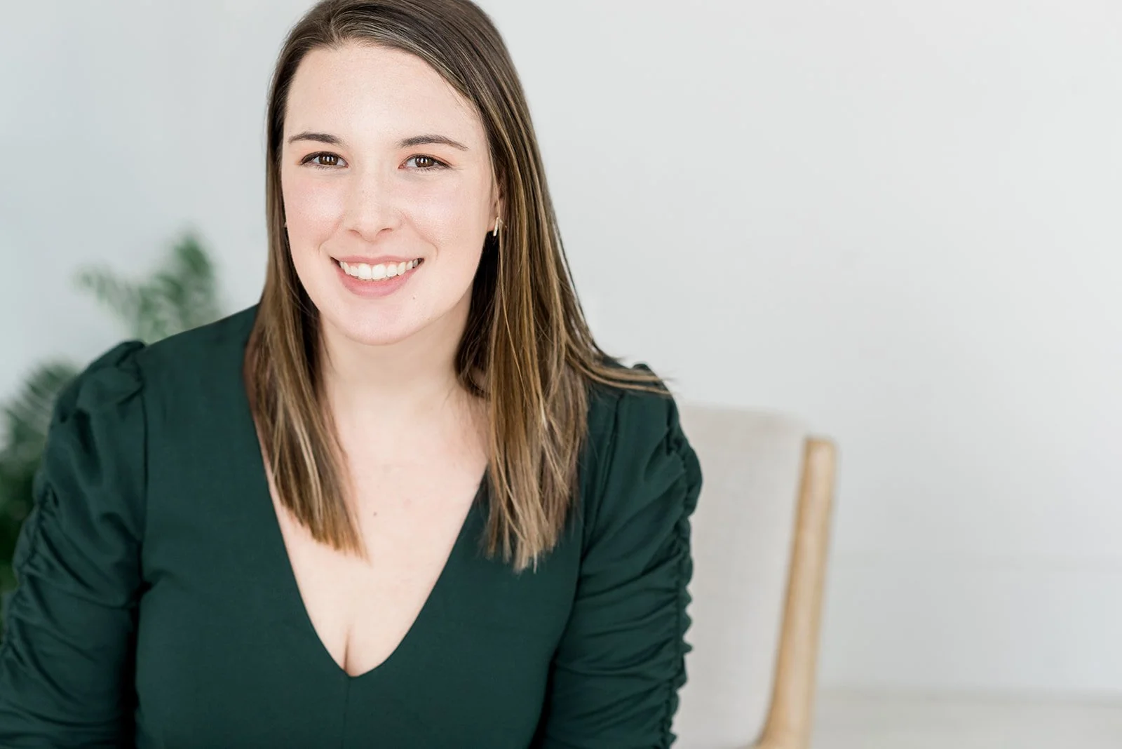A smiling woman with brown hair wearing a dark green top, sitting indoors with a white wall and a part of a plant in the background.