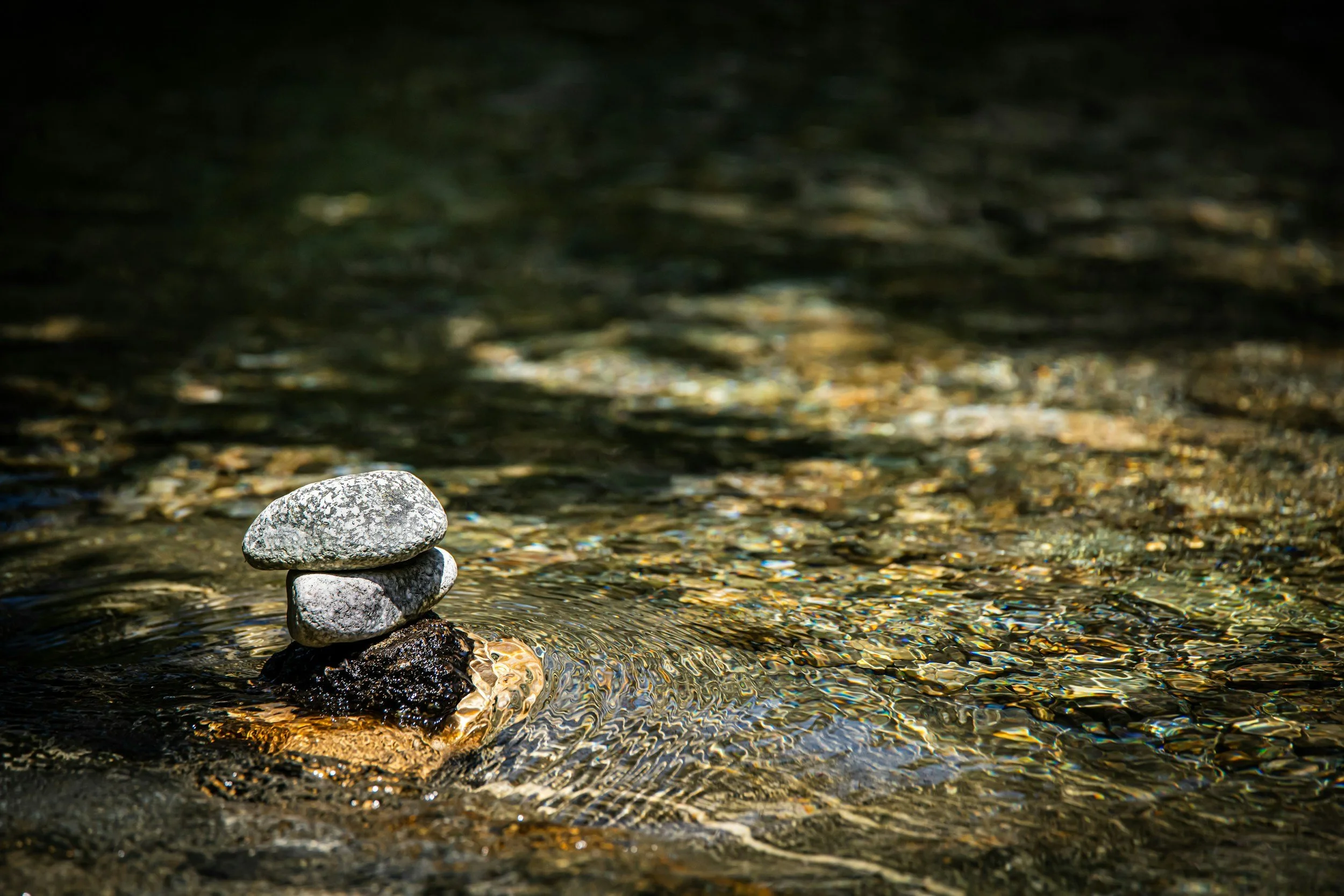 A small stack of three gray stones balanced on top of a dark rock in a shallow, clear stream with rippling water.