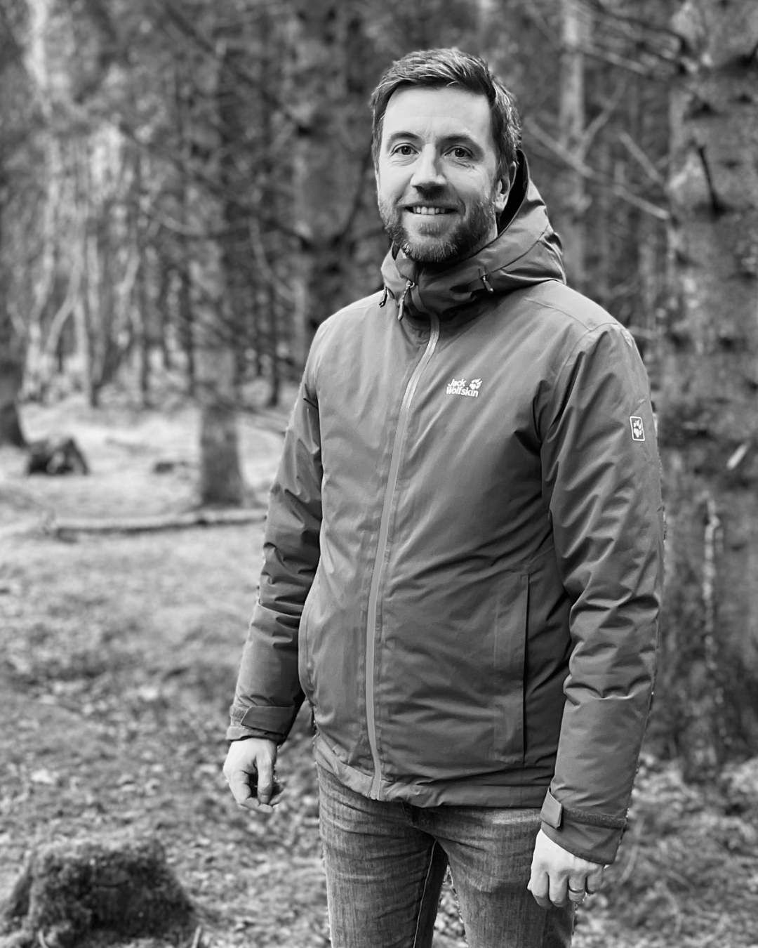 A man standing outdoors in a wooded area, wearing a jacket and smiling.