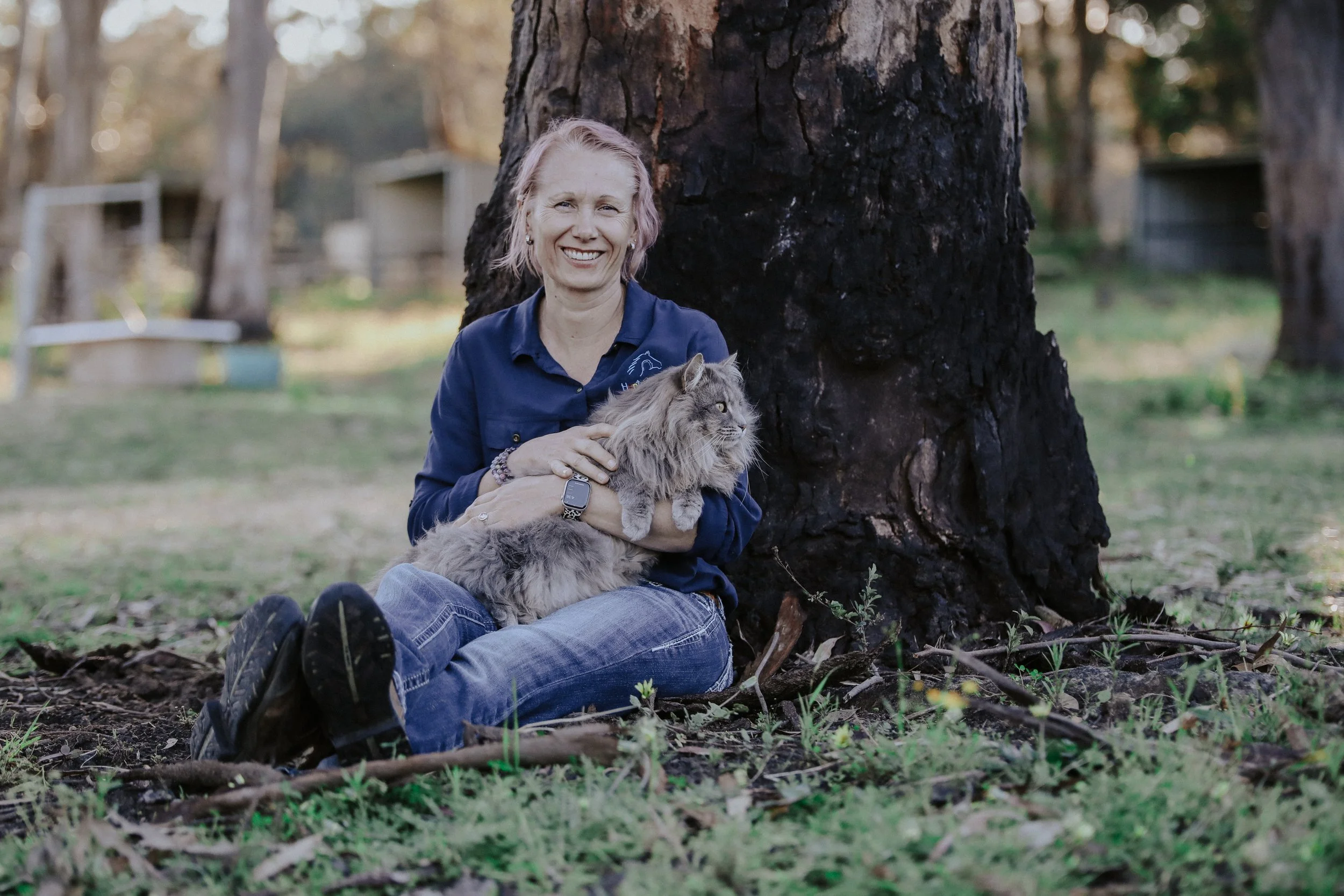 A woman sitting on the ground next to a large tree trunk, holding a fluffy gray cat, outdoors in a natural setting with trees and a fence in the background.