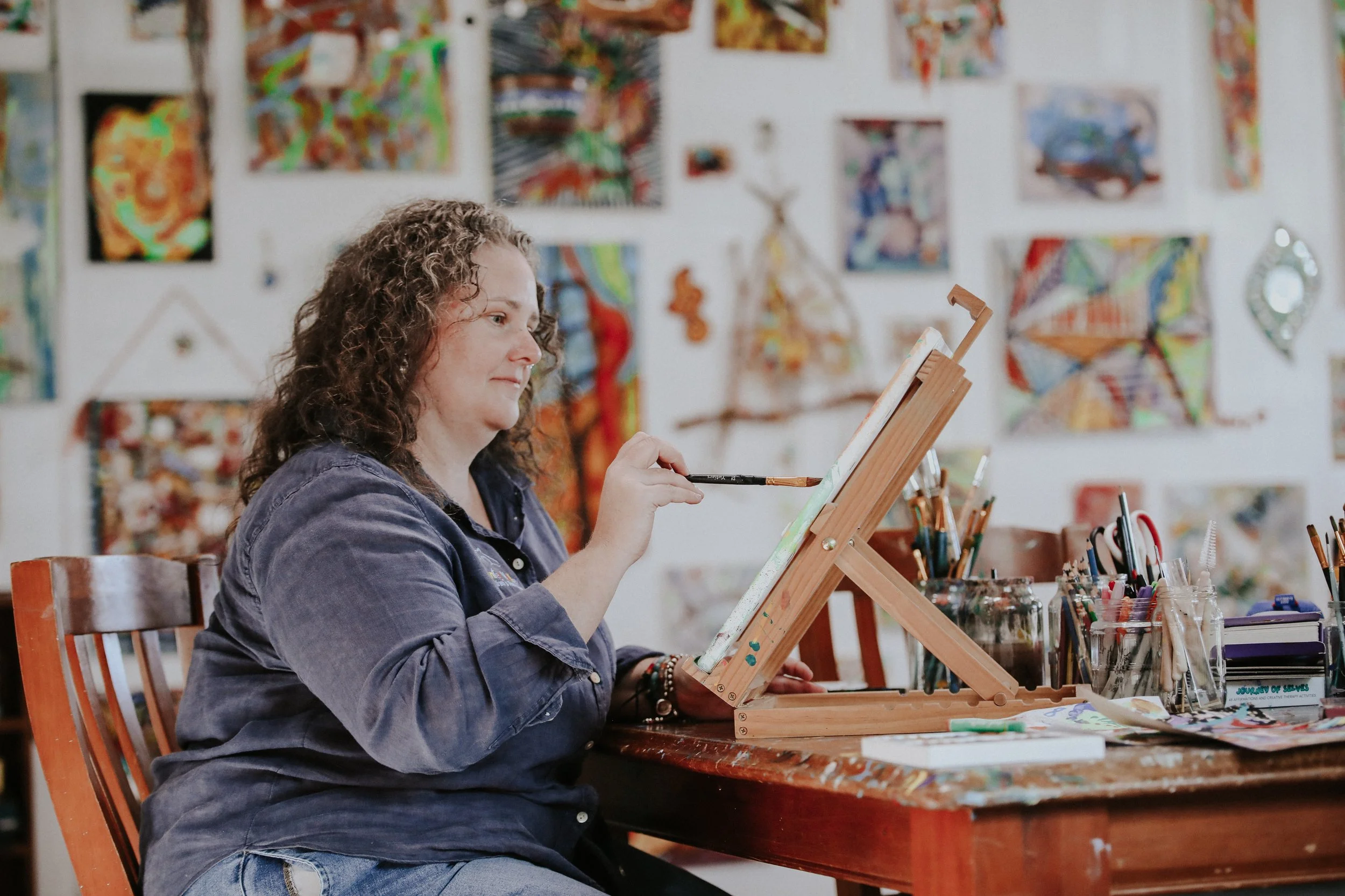 A woman with curly hair sits at a wooden table, painting on a small canvas while surrounded by art supplies in an art studio. The background features colorful abstract paintings and artworks on the wall.
