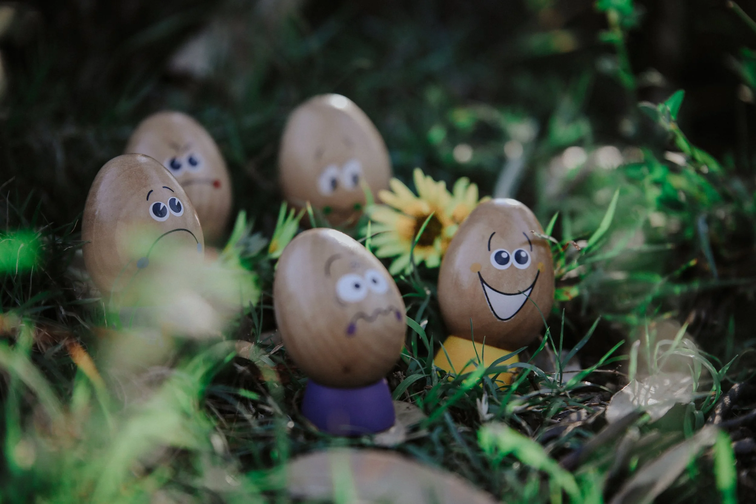 Several decorated eggs with painted faces and expressions, positioned among grass and small plants, with a yellow flower in the background.