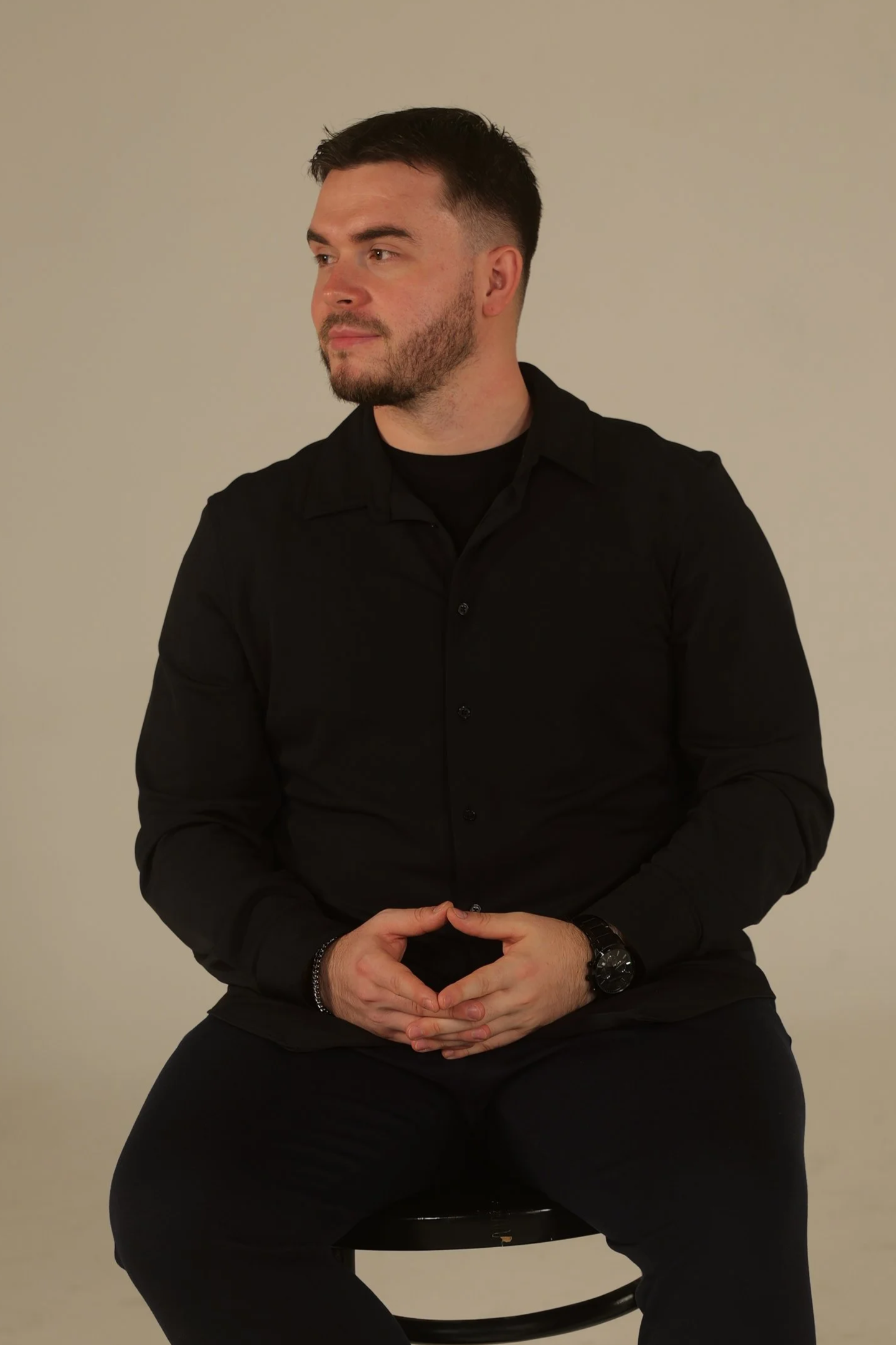 A man sitting on a stool against a plain light-colored background, wearing a black shirt and black pants, with short dark hair and a beard, looking to the side with hands clasped in his lap.
