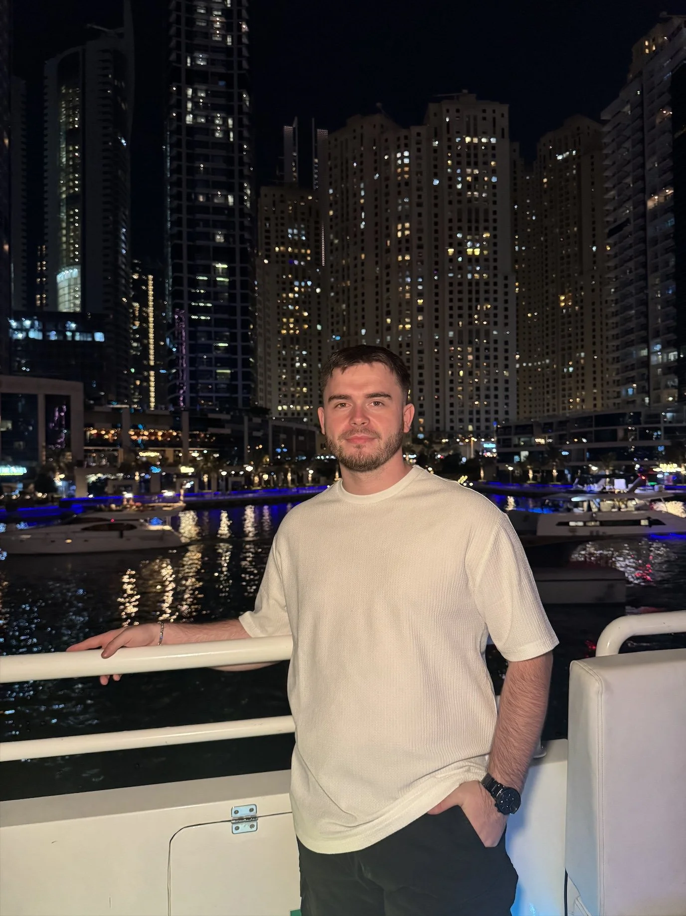 A man in a white shirt stands on a boat at night with a city skyline and marina in the background.