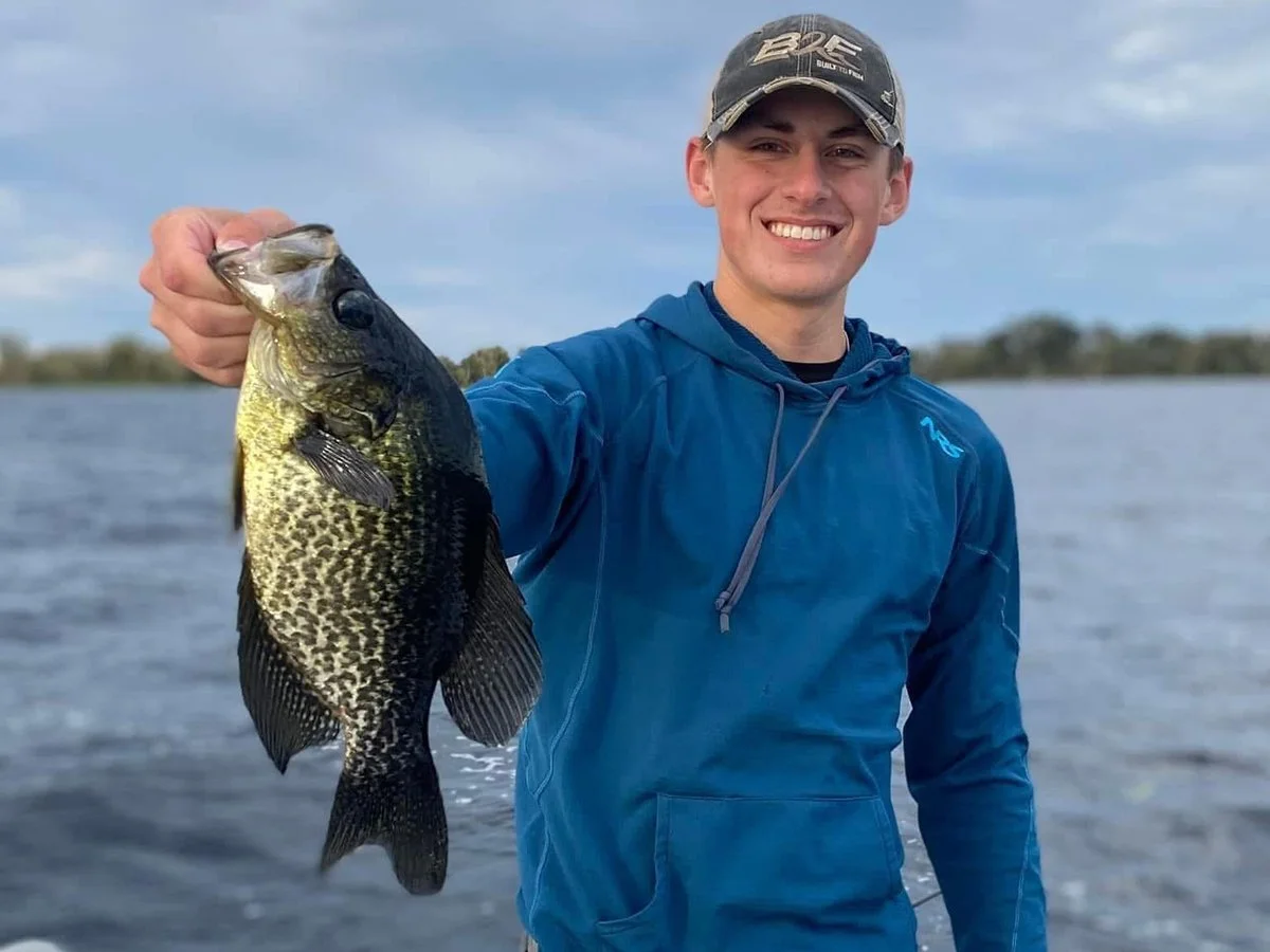 A young man in a blue jacket and baseball cap smiling while holding a large fish in front of a body of water during daytime.