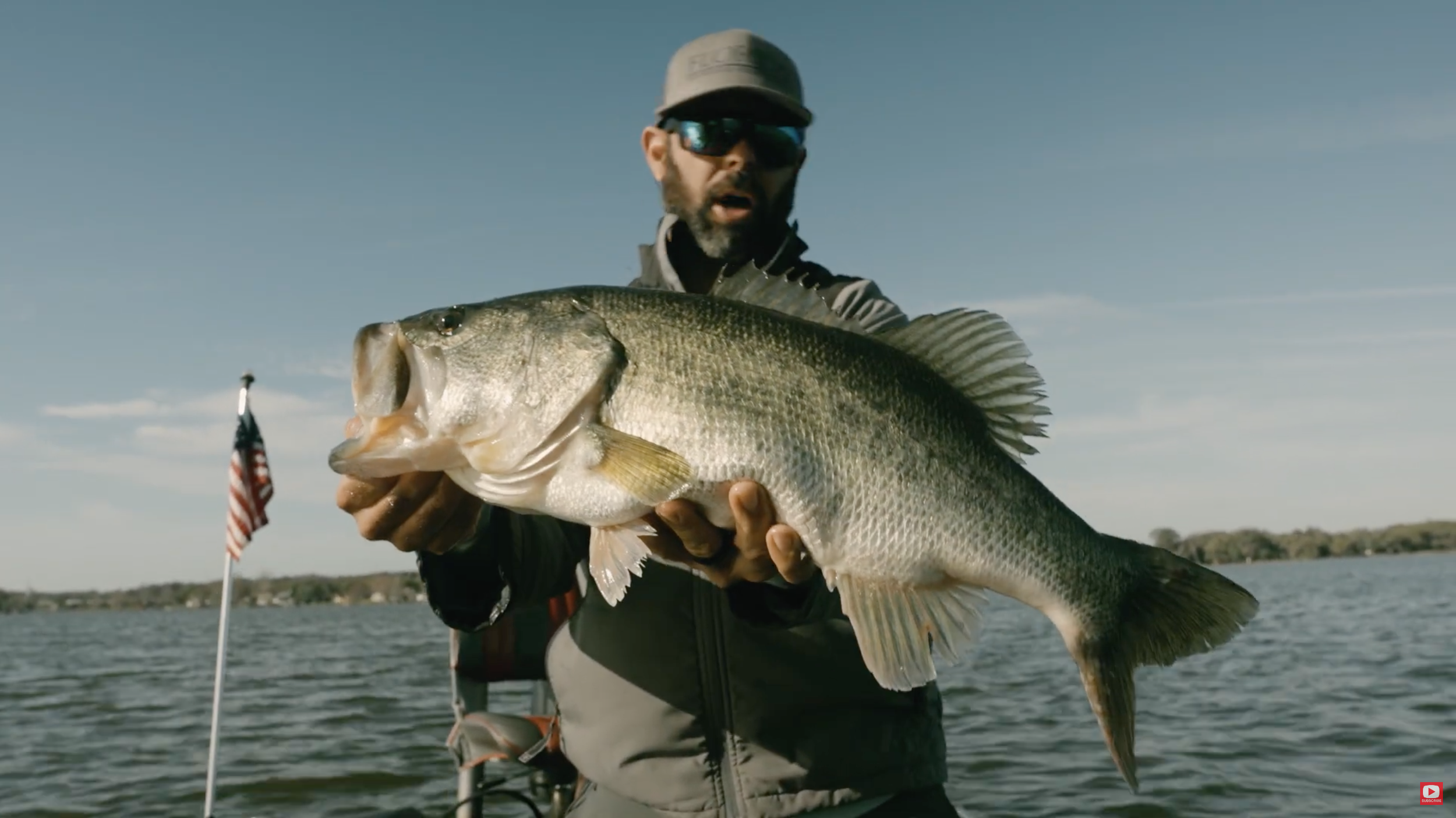 Man in sunglasses and cap holding a large fish on a boat with water and sky background.