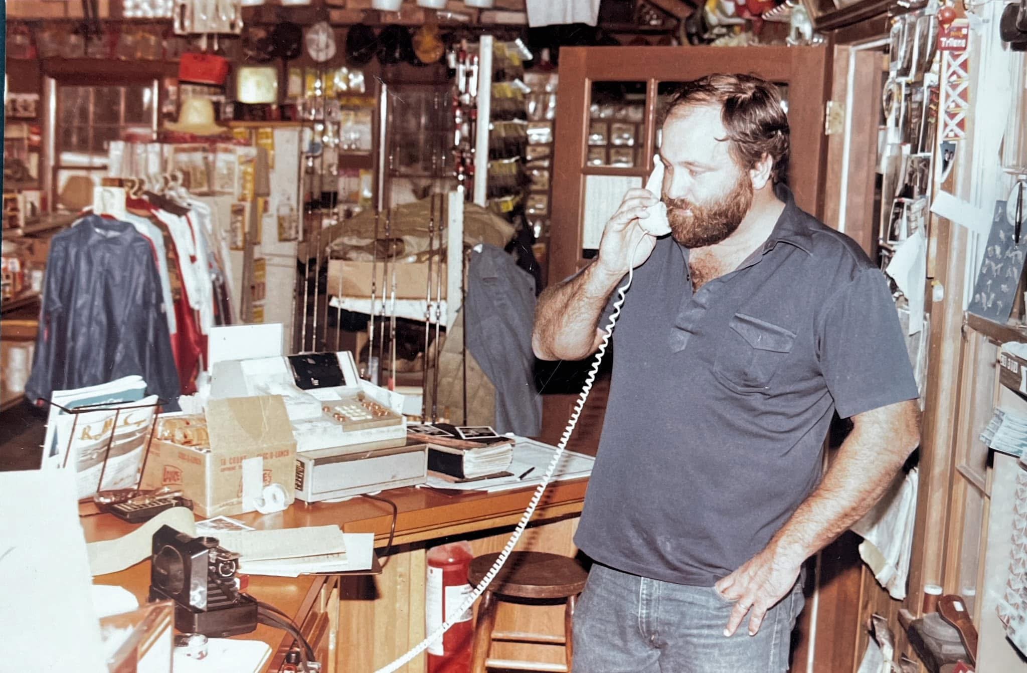 A man with a beard and casual clothing is talking on the telephone inside a cluttered hardware or general store, surrounded by various tools, clothes, and items on shelves and counters.