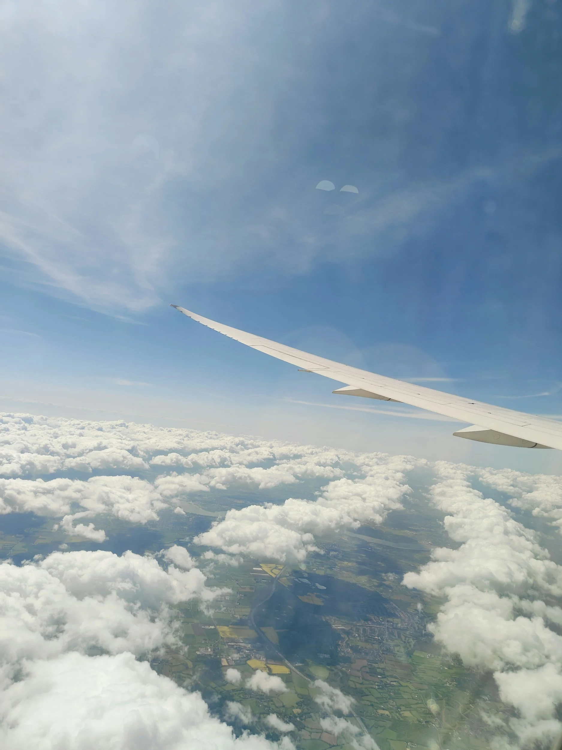 View from an airplane window showing the wing of the plane and a landscape of clouds, fields, and a river below, with a partly cloudy blue sky.