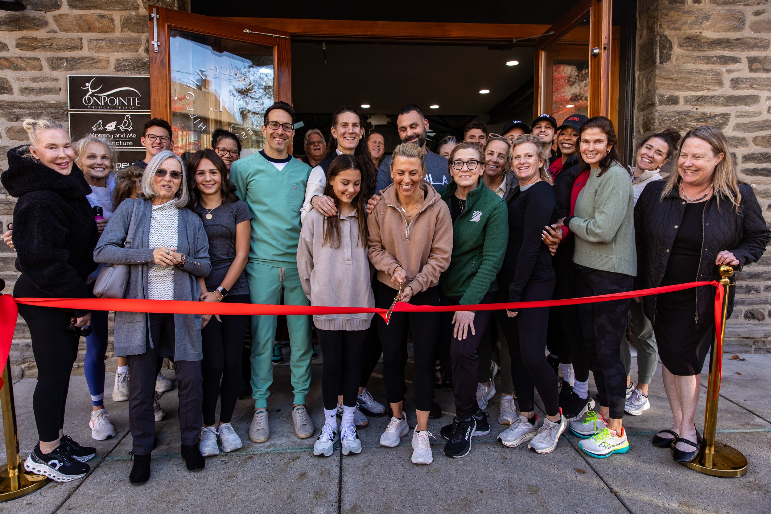 Group of people at a ribbon-cutting ceremony outside a brick building, smiling and gathered around a woman cutting a red ribbon.