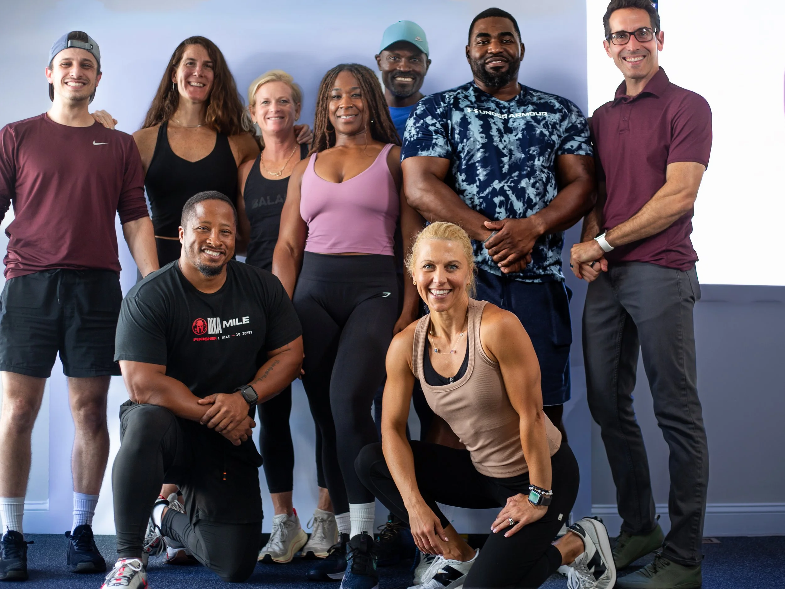 Group of ten people, diverse in ethnicity and gender, posing indoors against a plain wall, dressed in athletic clothing, smiling at the camera.