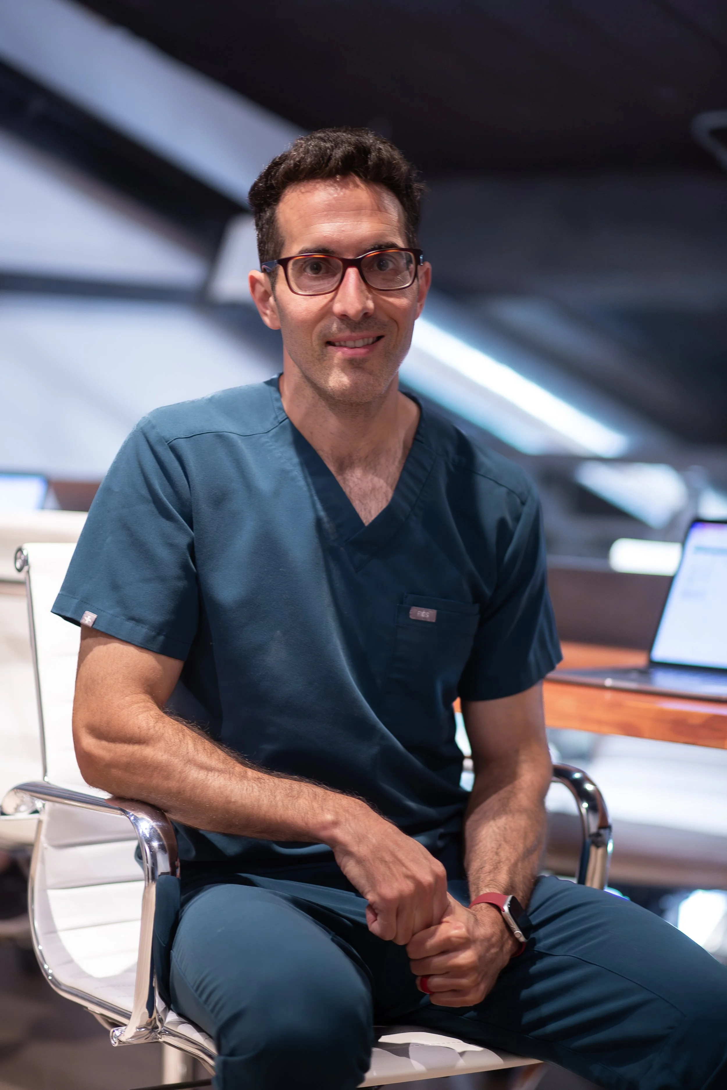 A man in medical scrubs sitting on a white chair, smiling, in a modern office or medical environment.