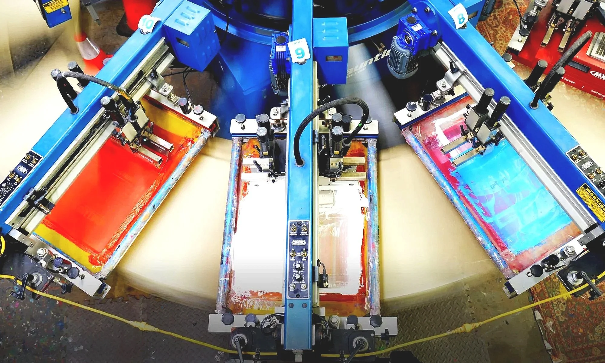 A screen printing press in use with three screens, color ink, and various mechanical parts.
