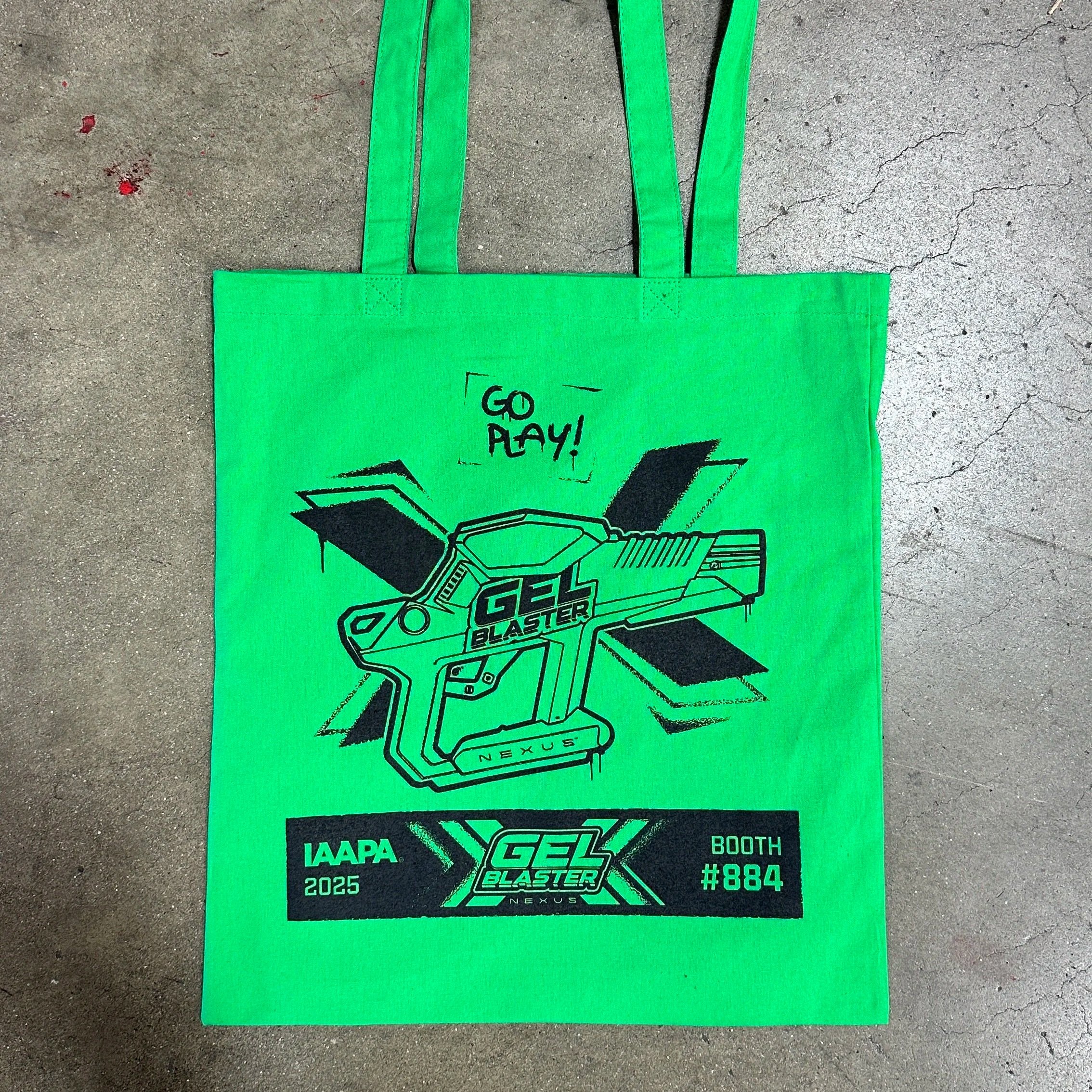 A bright green tote bag with black graphics and text for the IAPA 2025 event. Features a drawing of a futuristic toy gun labeled "GEL BLASTER" with wings and the text "GO PLAY!" above. The bottom section has event information: "IAPA 2025," event name "GEL BLASTER," a booth number "#884"