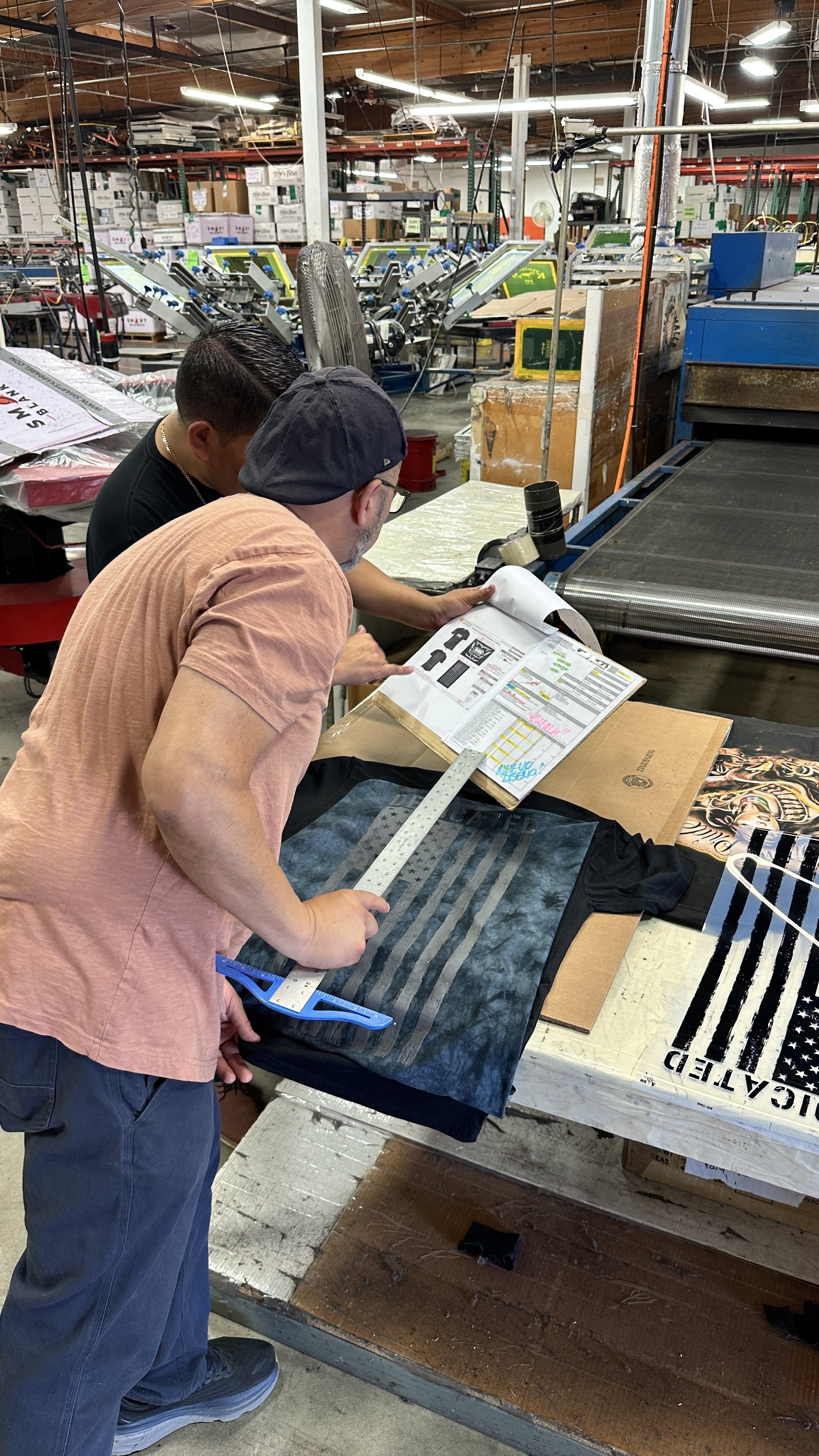 Two people are working on a printing project in an industrial workspace with various printing machines and supplies in the background. One person is holding a ruler and a printout, examining a black fabric with a faded American flag design.