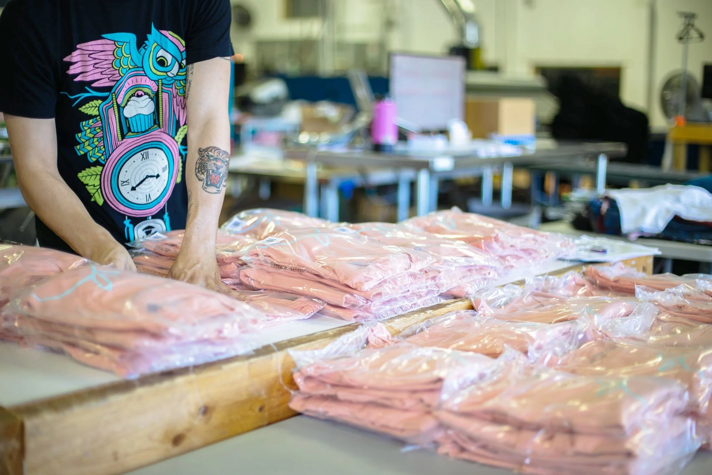 Person wearing a black t-shirt with colorful graphic design, including a clock and a tiger tattoo, handling pink vacuum-sealed packages of meat on a worktable in a meat processing or packaging facility. Background includes work tables, computers, and industrial equipment.
