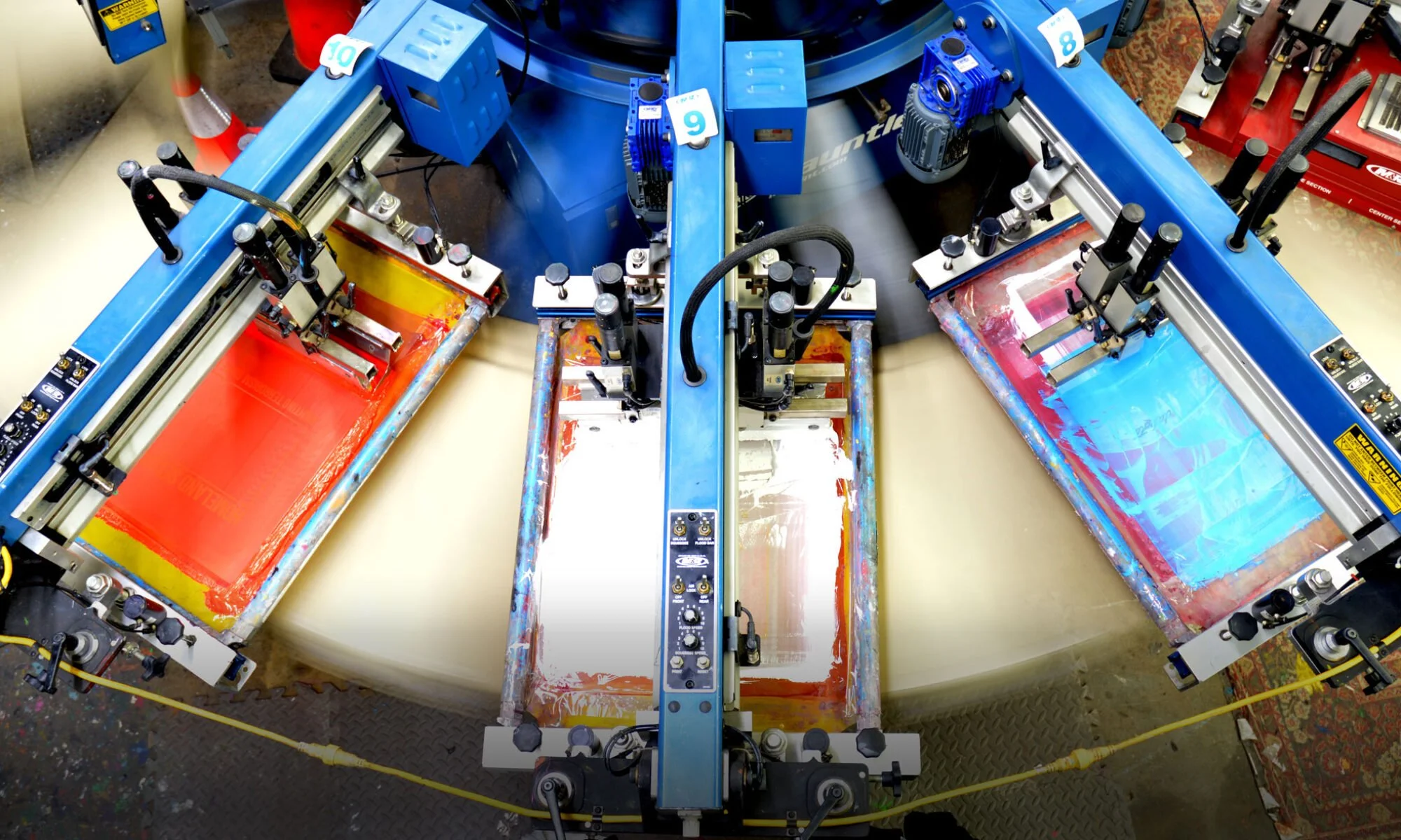 Close-up of three screen printing presses with squeegees and screens, used for applying ink onto fabrics or other materials.
