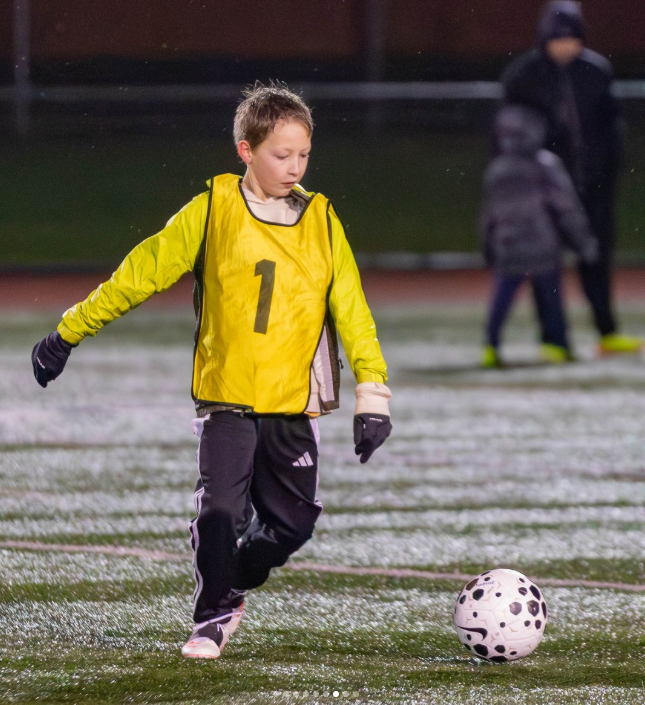 Young boy in yellow sports vest with number 1, black gloves, black pants, and white shoes dribbling a black and white soccer ball on a wet outdoor field at night.
