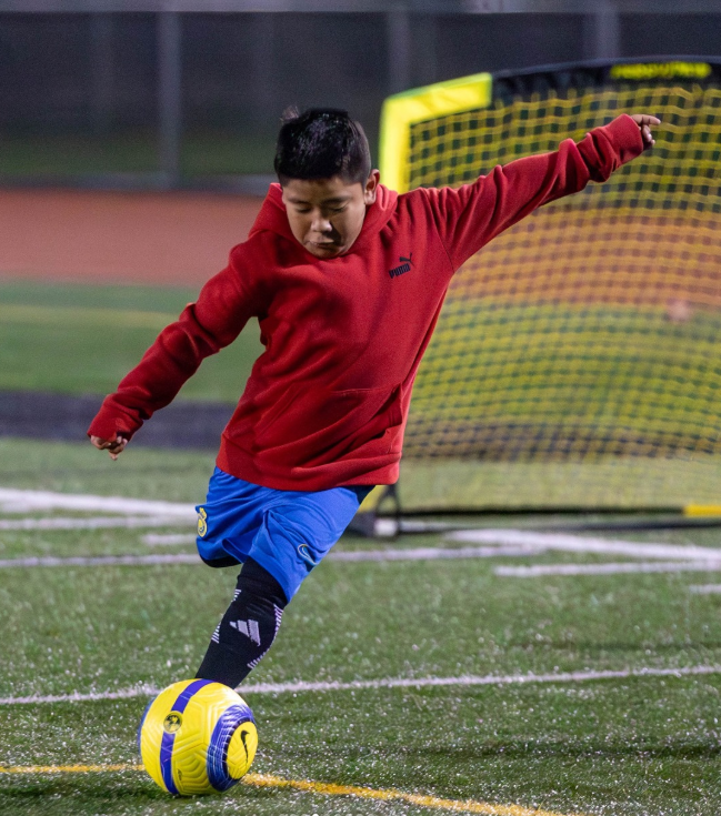 A young boy in a red hoodie and blue shorts playing soccer outdoors on a field with artificial turf, kicking a yellow and blue soccer ball near a yellow goal net in the background.