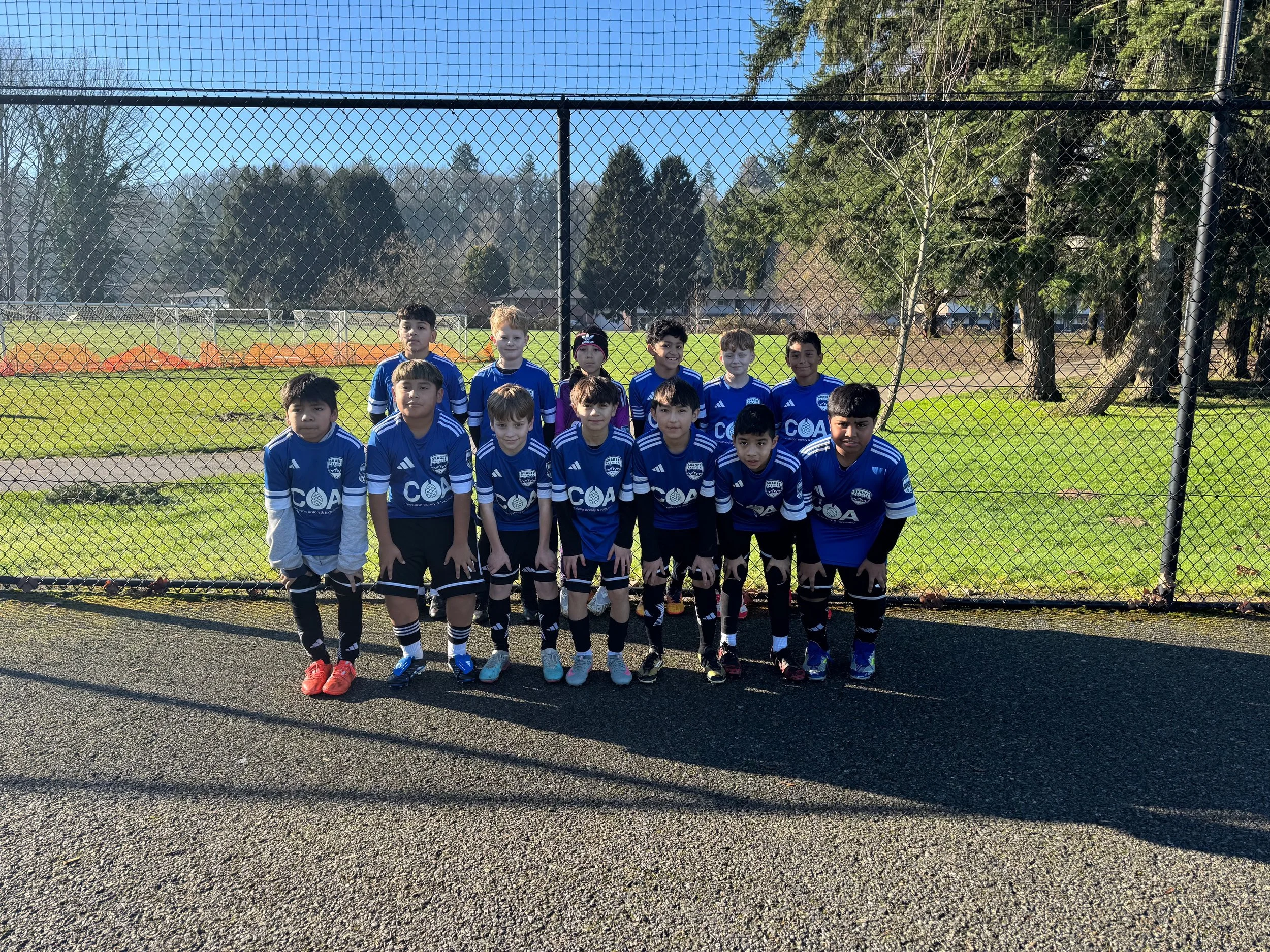 A group of young boys in blue soccer jerseys posing in front of a chain-link fence, outdoors on a sunny day, with a grassy field and trees in the background.