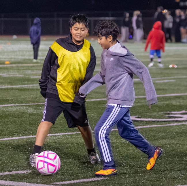 Two boys playing soccer on a field at night, one wearing a yellow vest over a black shirt and the other in a gray hoodie and blue pants, with a pink and white soccer ball between them.