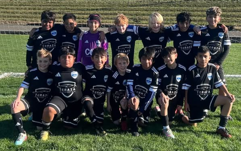A youth soccer team of twelve kids, dressed in black uniforms with a blue and white emblem and sponsor logo, posing for a group photo on a grassy field. They are standing in front of a soccer goal, with some kneeling in the front row and others standing behind them.
