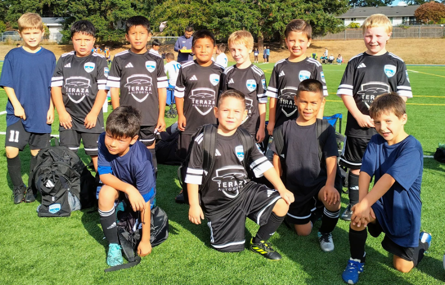 Young boys on a soccer field posing for a team photo, wearing black and blue soccer uniforms and cleats, with a few carrying backpacks.