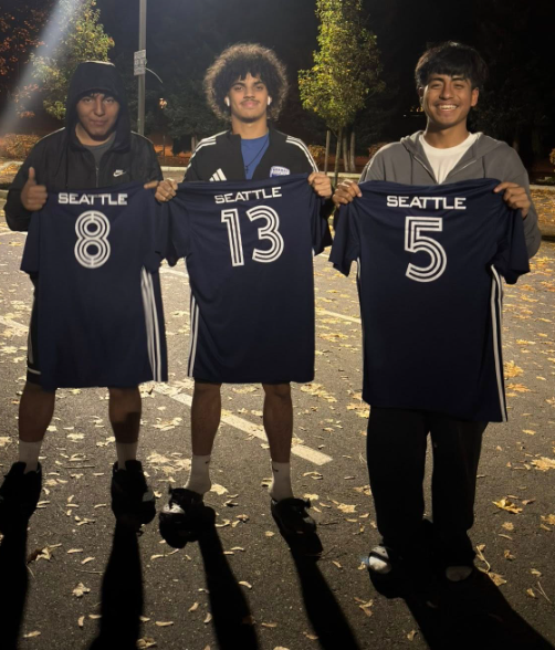 Three young men holding Seattle soccer jerseys with numbers 8, 13, and 5, outdoors at night, surrounded by trees with autumn leaves on the ground, smiling and posing for the photo.