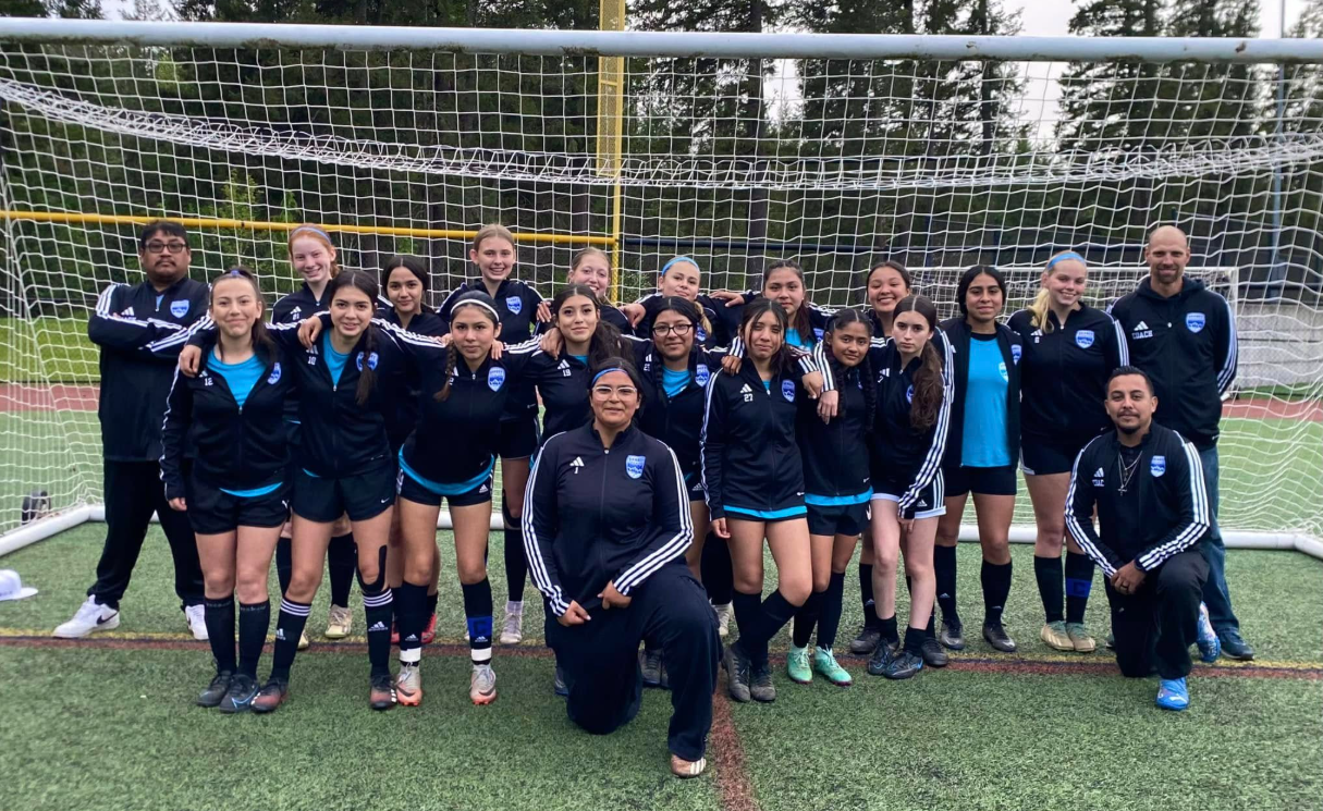 Group of young female soccer players and coaches on a field, posing in front of a goalpost