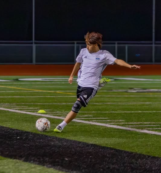 A young boy wearing a white soccer jersey and black shorts kicking a soccer ball on a green turf field at night.
