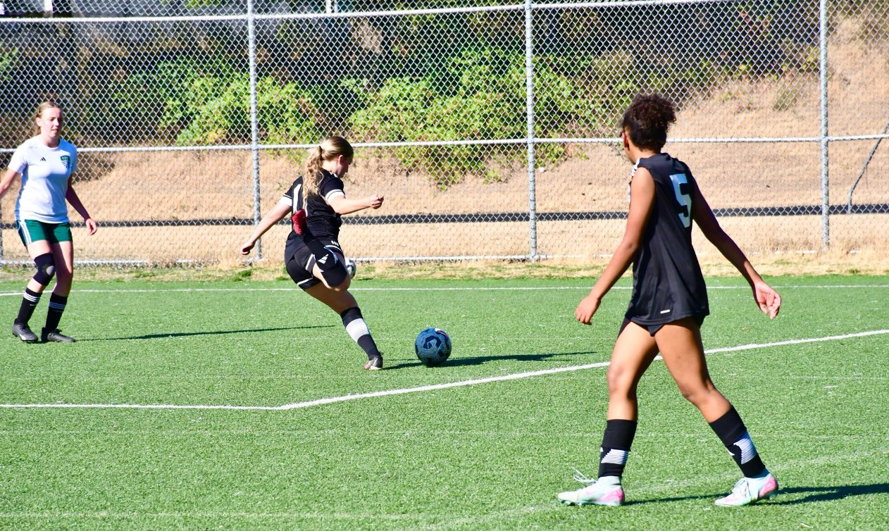 Three young women playing soccer on a green field, one kicking the ball, and two watching nearby.